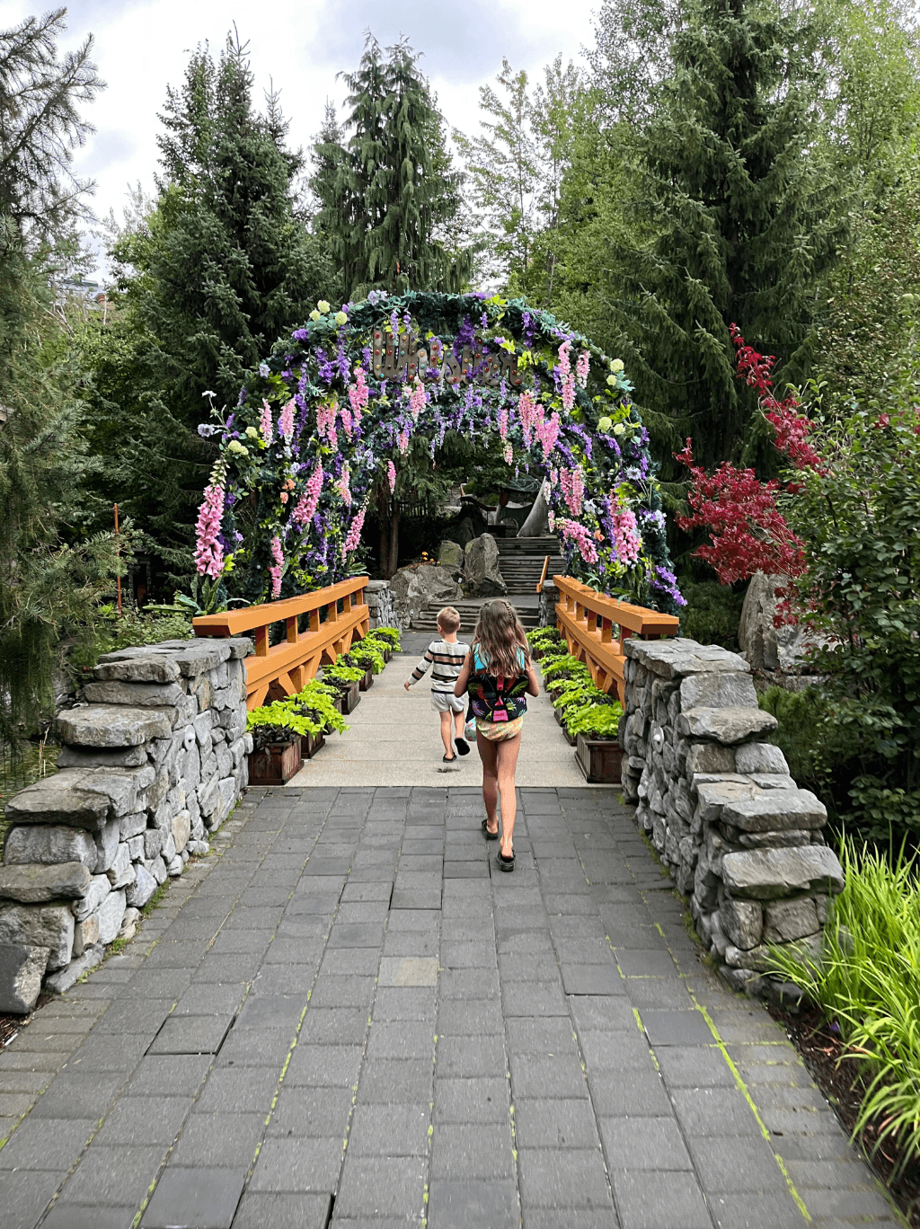Two children walking under a floral archway surrounded by pine trees and stone.