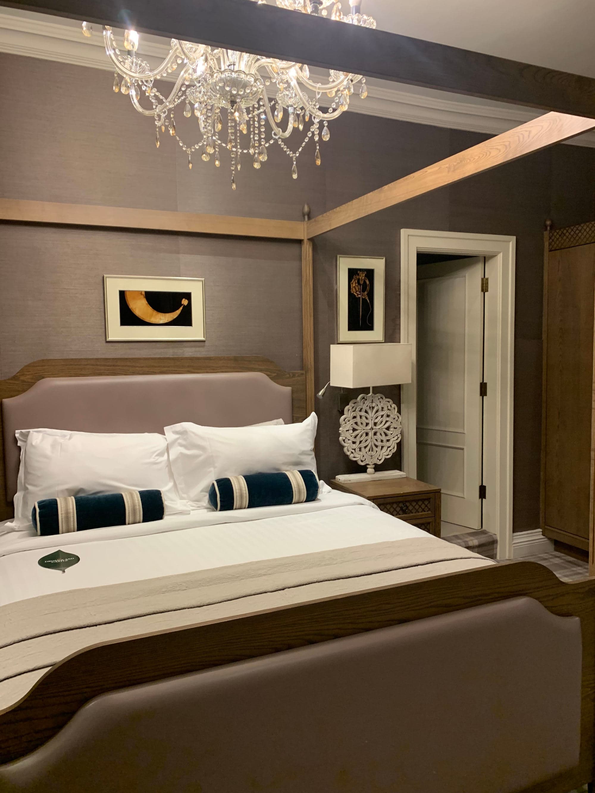 A bedroom with decorative pillows, artwork, lamps and a fancy chandelier hanging above.