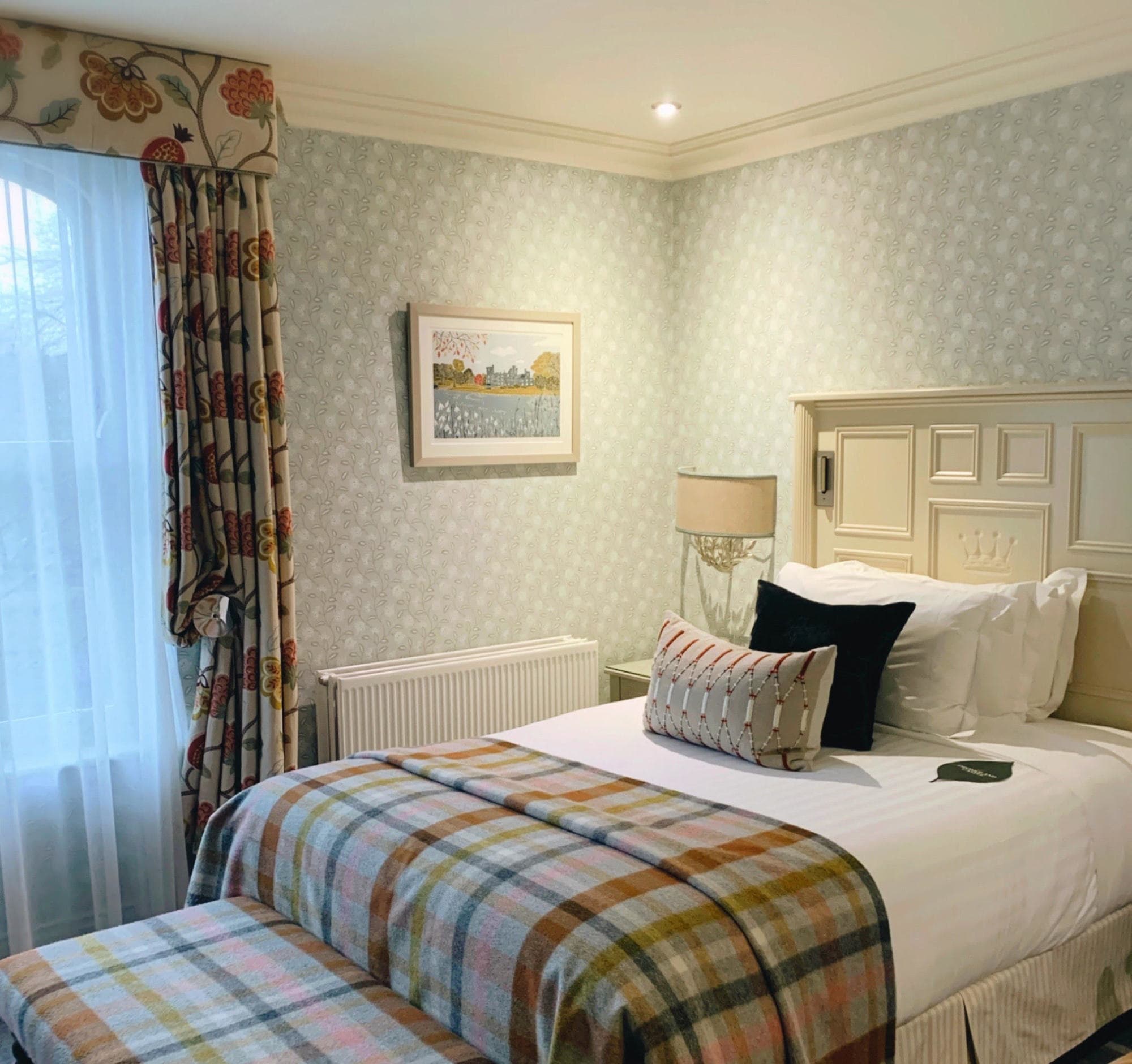 A bedroom with wallpaper, decorative blankets and pillows, art and curtains.