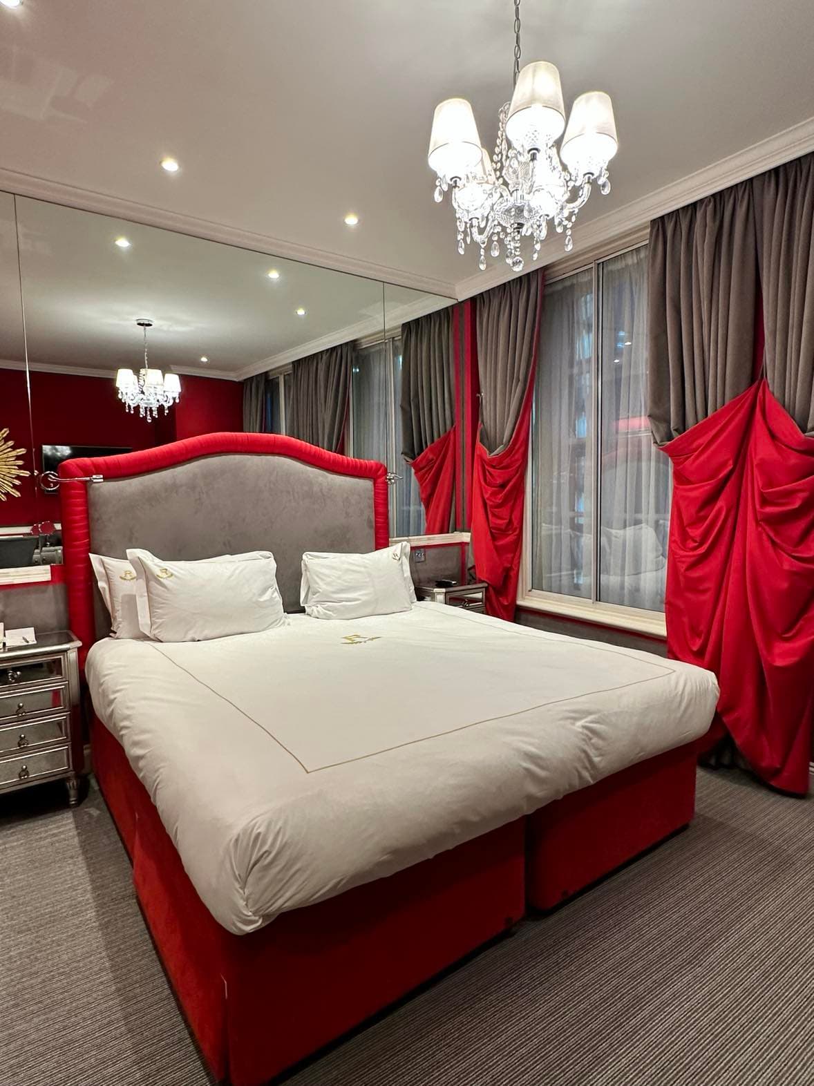 A view of the bedroom complete with a red lined headboard, white bedding, white chandelier, mirrored wall and red and grey curtains on the window.