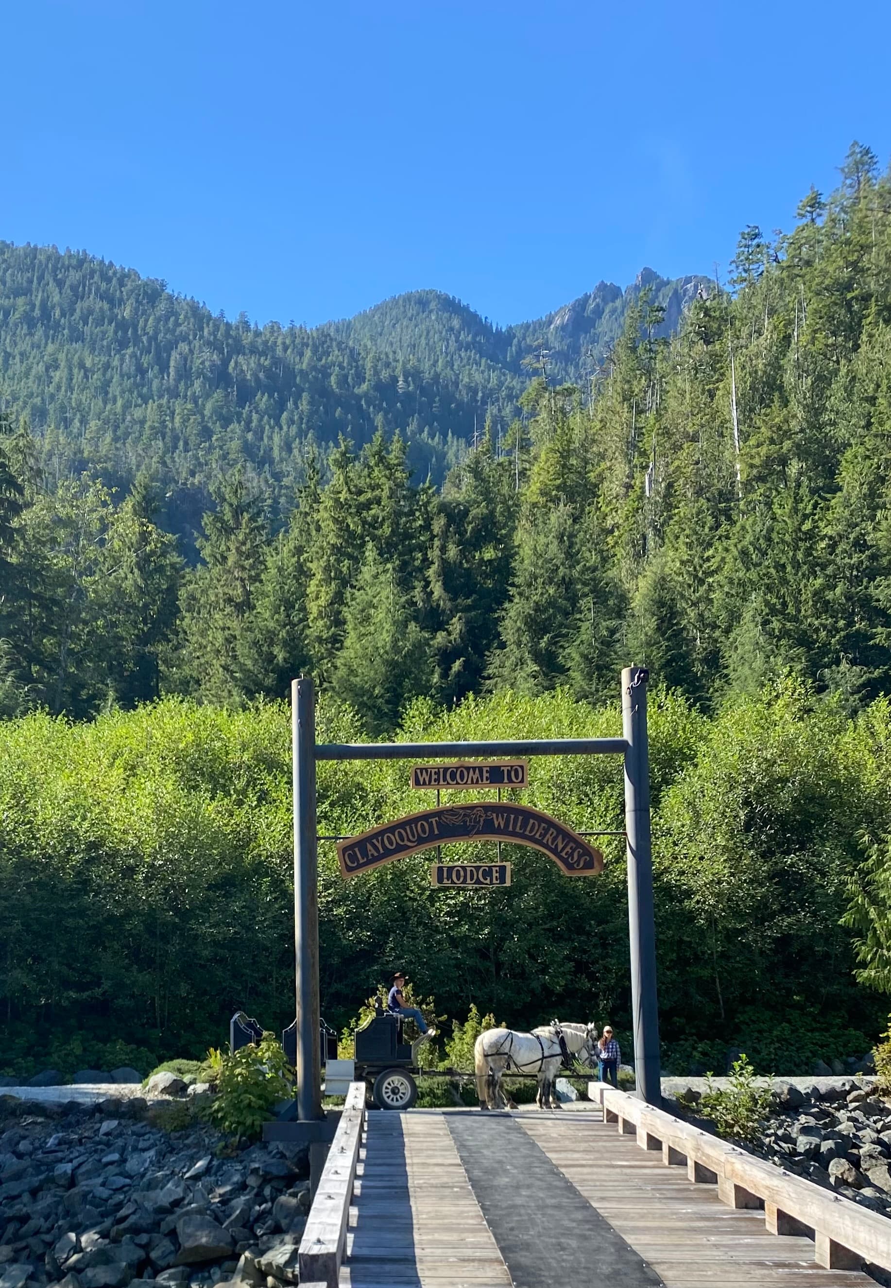 A wooden bridge leading to an archway with a sign in front of a view of trees and a large green mountain.
