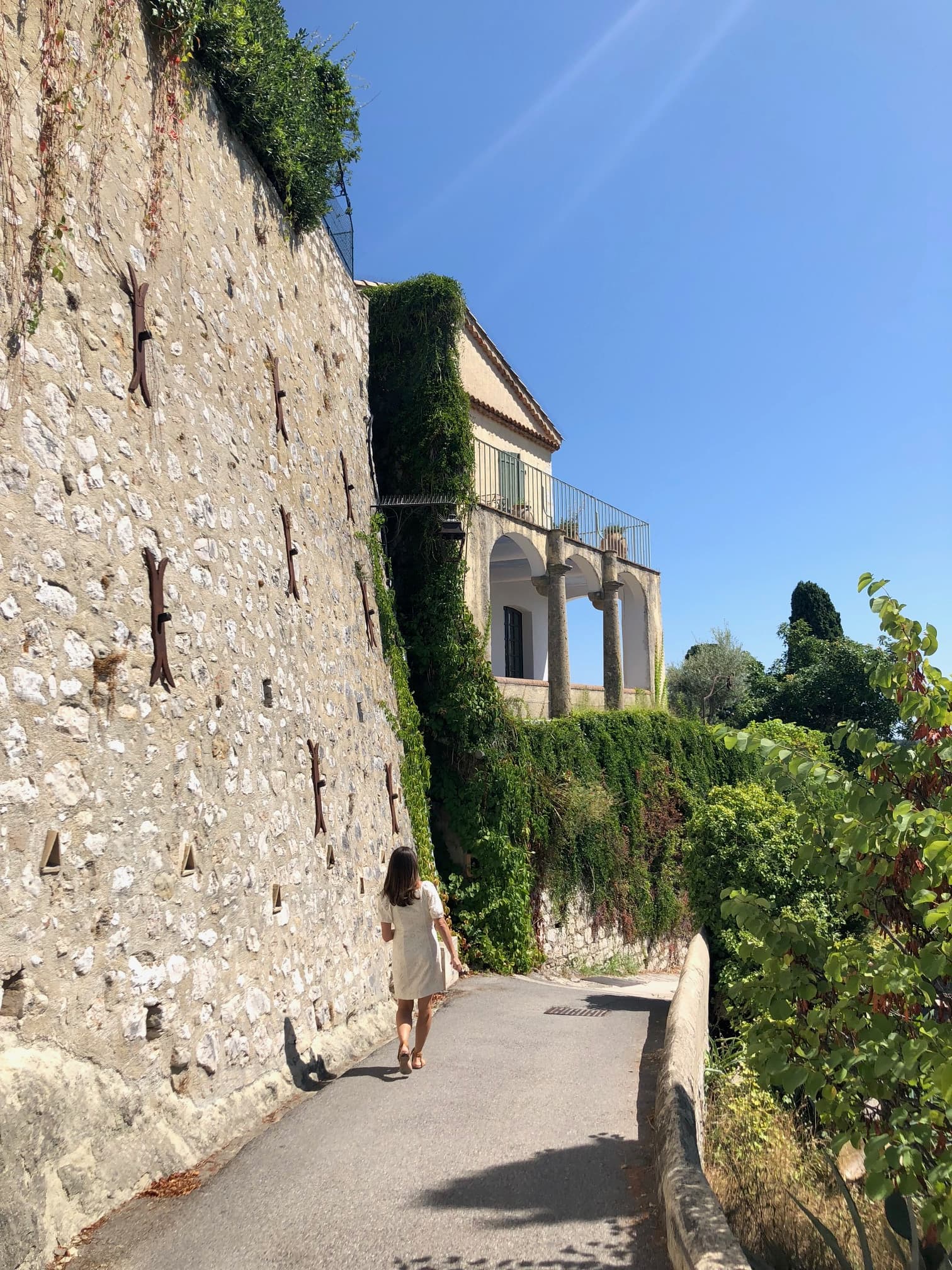 A woman walking along a road next to a stone wall with a terraced house and greenery beyond