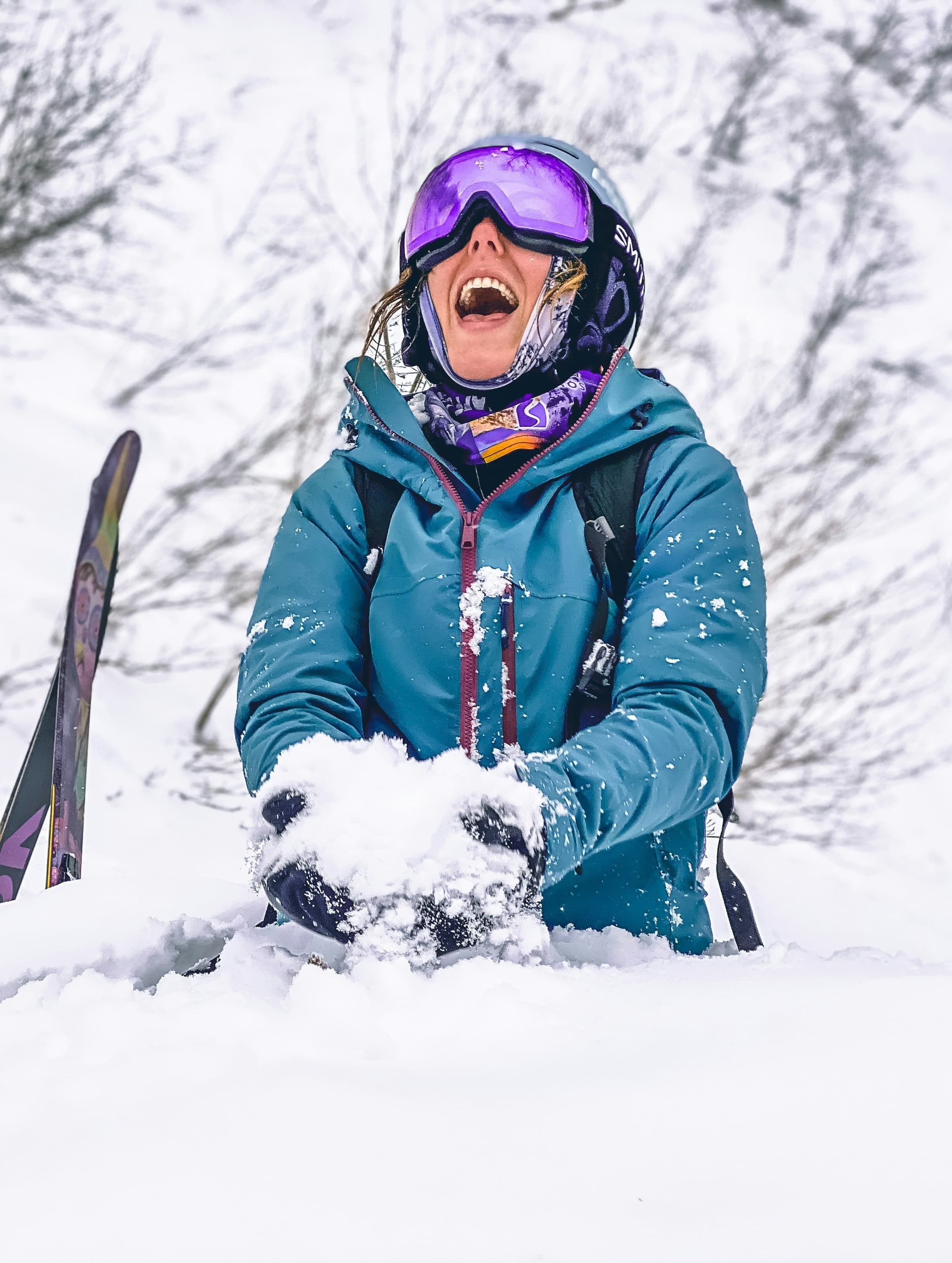 A person wearing a teal jacket and purple ski goggles sitting down in a pile of snow with their mouth smiling and wide open while holding snow in their hands.