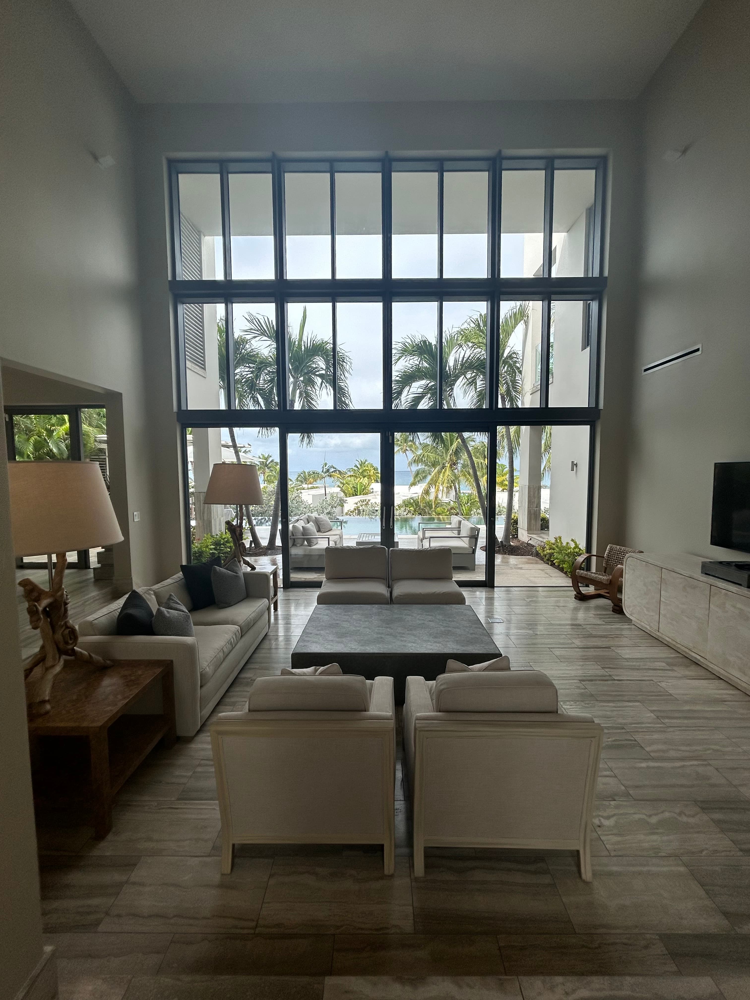 A beautiful view of a hotel suite complete with chairs, a couch, lamps, a table, a media console, a television and floor to ceiling windows with dark beams looking out to a view of palm trees and blue sky.