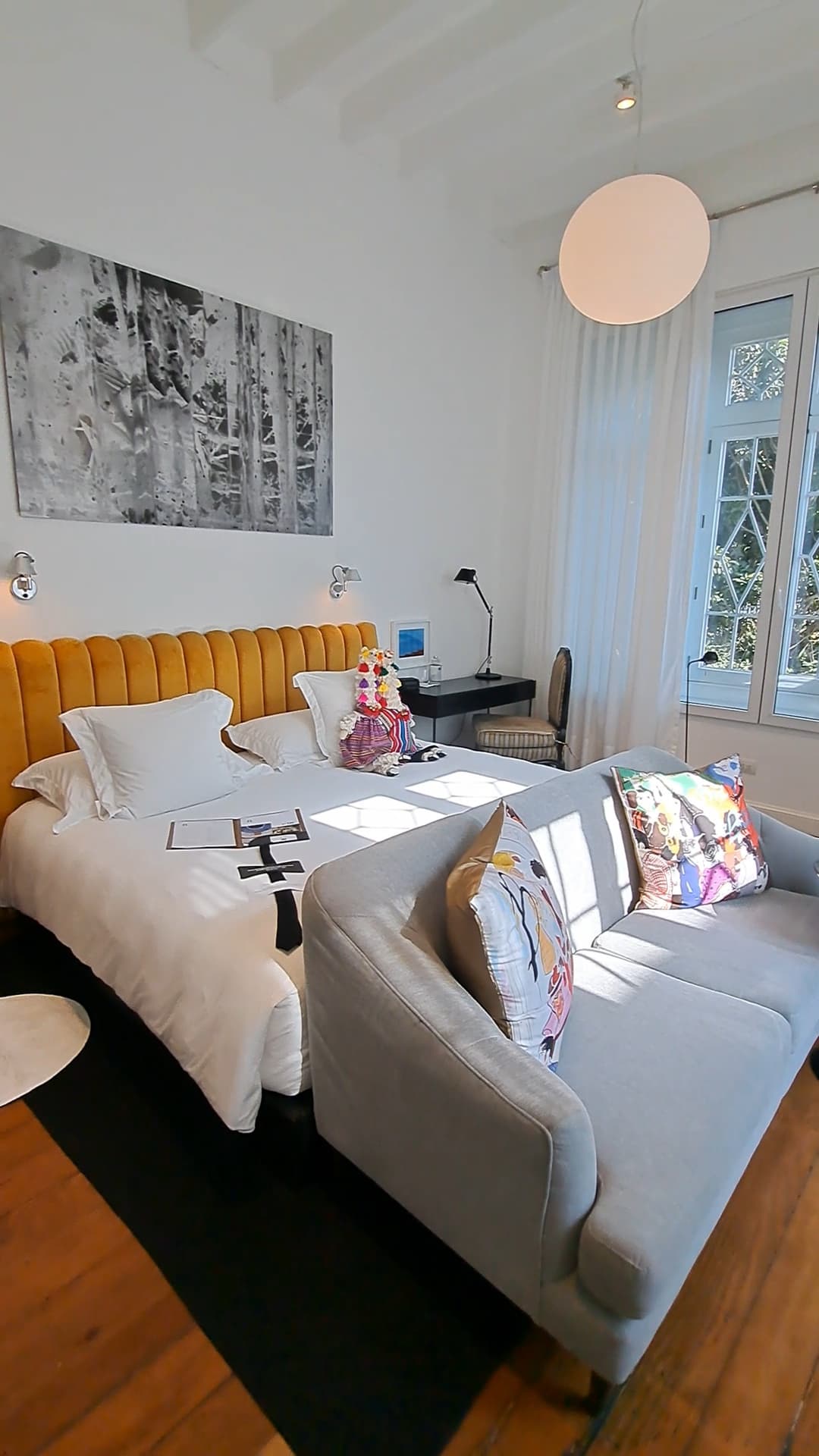 A bedroom with white bedding, a grey sofa at the foot of the bed, a mustard yellow headboard, artwork on the walls, decorative pillows and a round pendant light hanging from the white beamed ceiling.