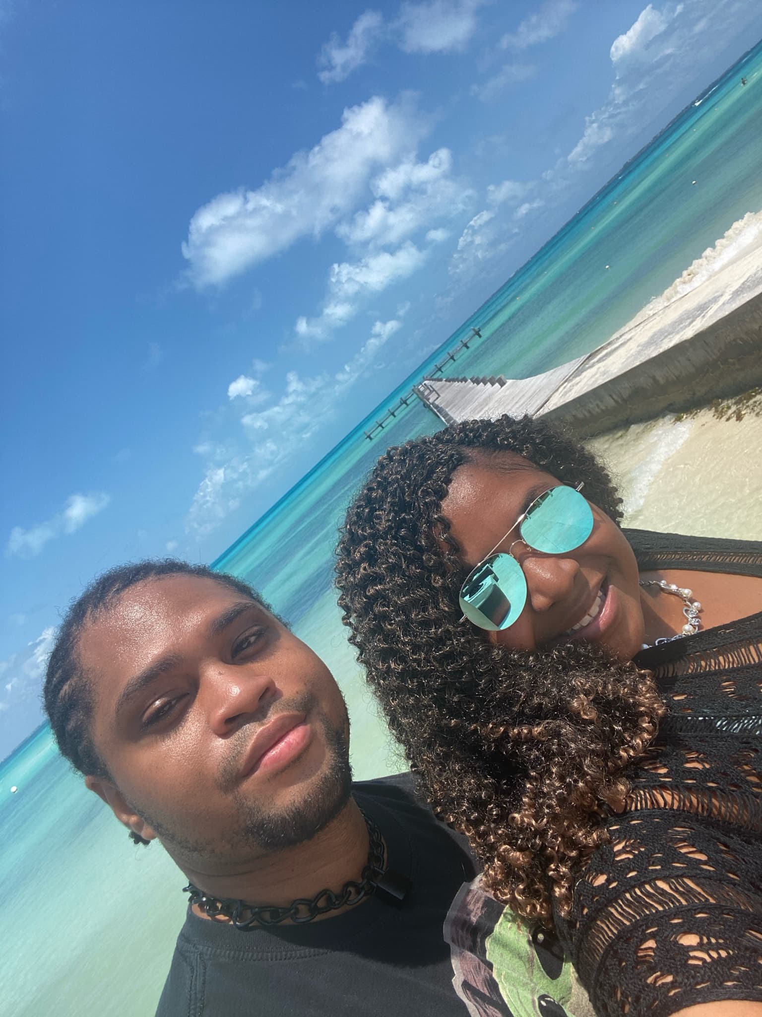 Two people posing for a selfie on the white sandy beach with the crystal-clear turquoise water in the background.