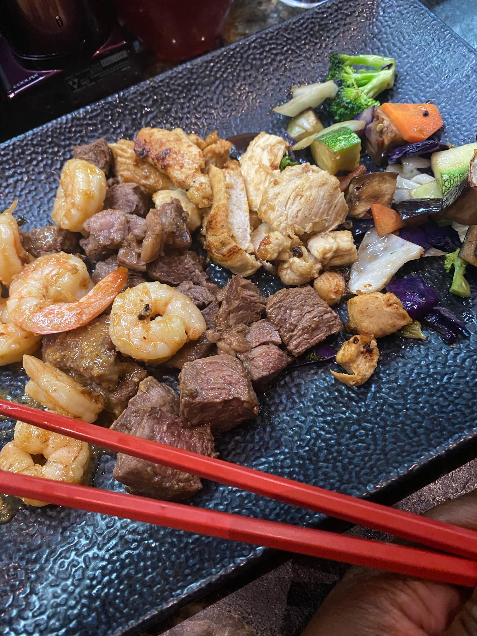 A dark plate full of various food items, such as shrimp, steak and vegetables. There are also red chopsticks being held above it.