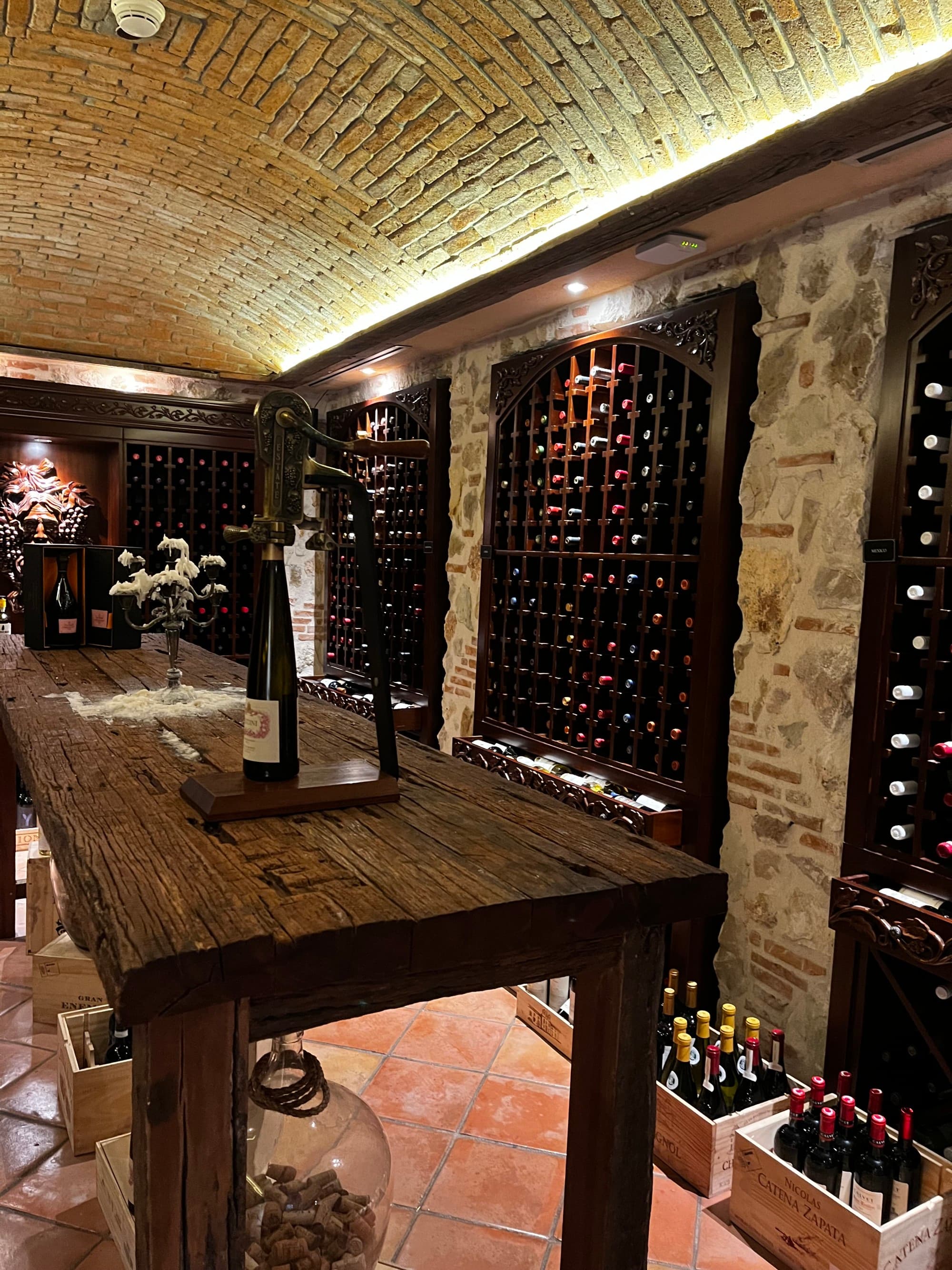 A beautiful stone wine cellar with wine bottles stored throughout it and a wooden table in the center of the room.