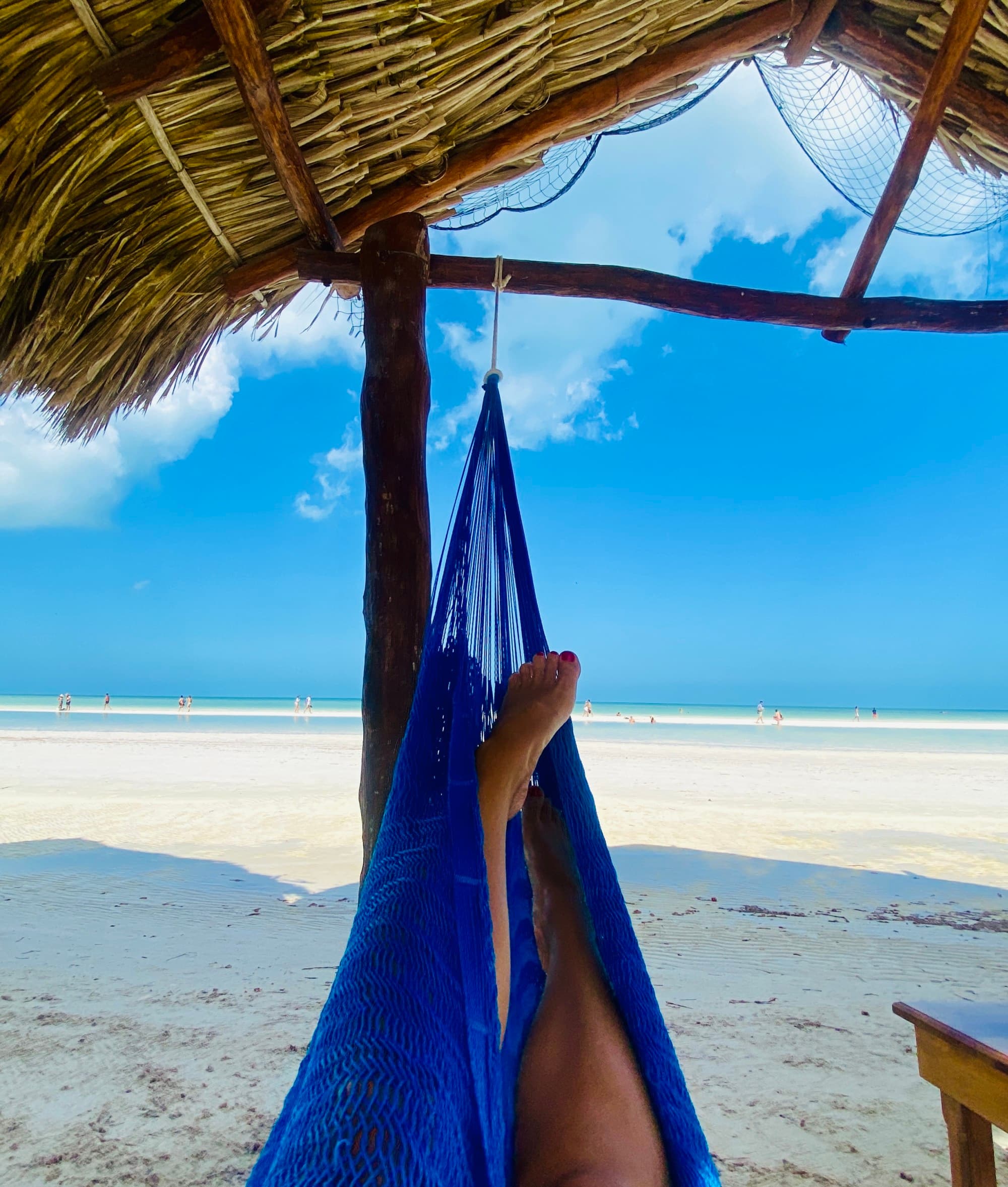 Two legs outstretched on a blue hammock outside as if a person is lounging. There is a straw hut roof above them and a sandy ground underneath. The ocean is in the far distance under the blue sky.