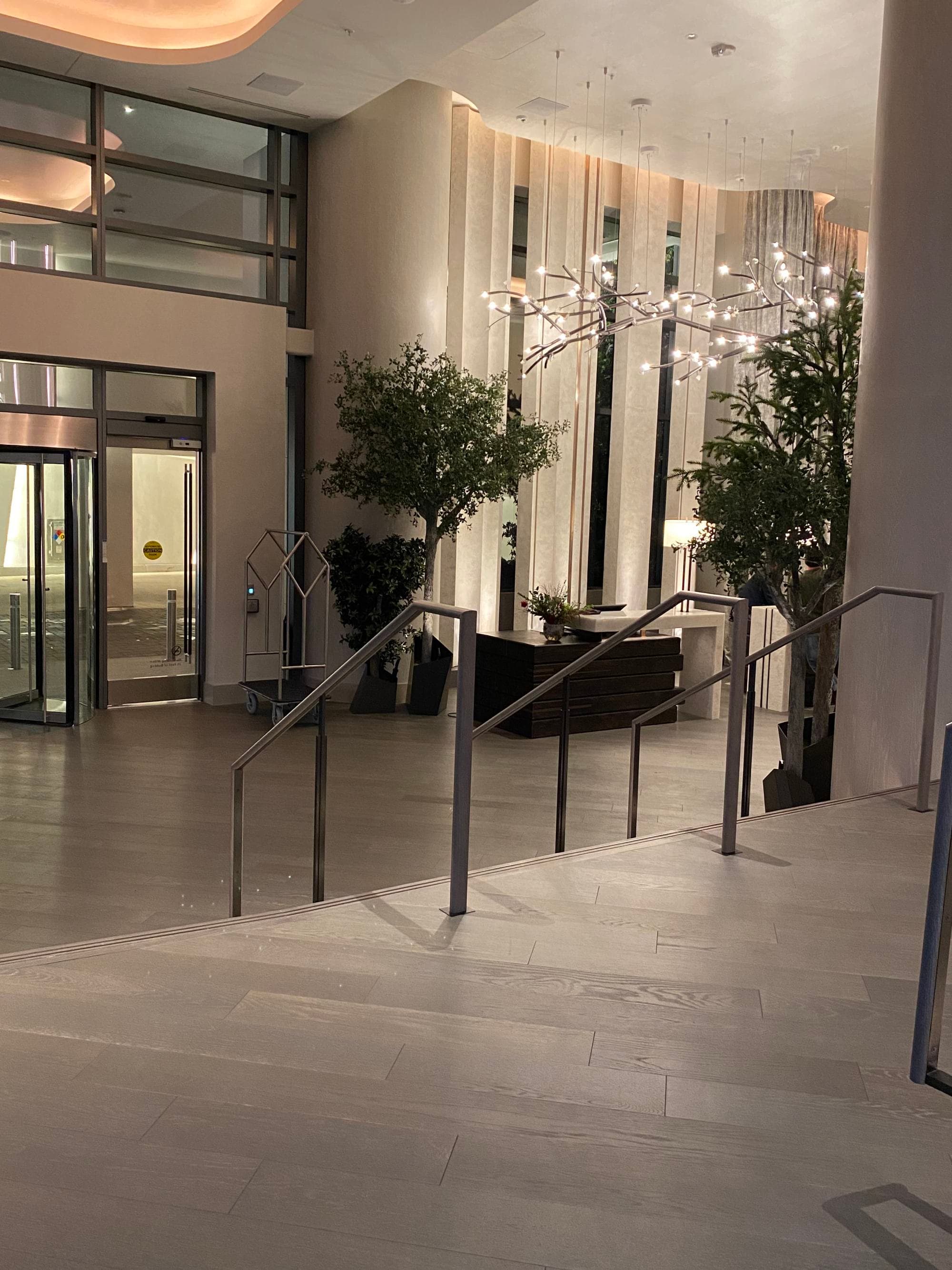 A hotel lobby with railings, chandeliers and white curtains. There are potted trees in the background.