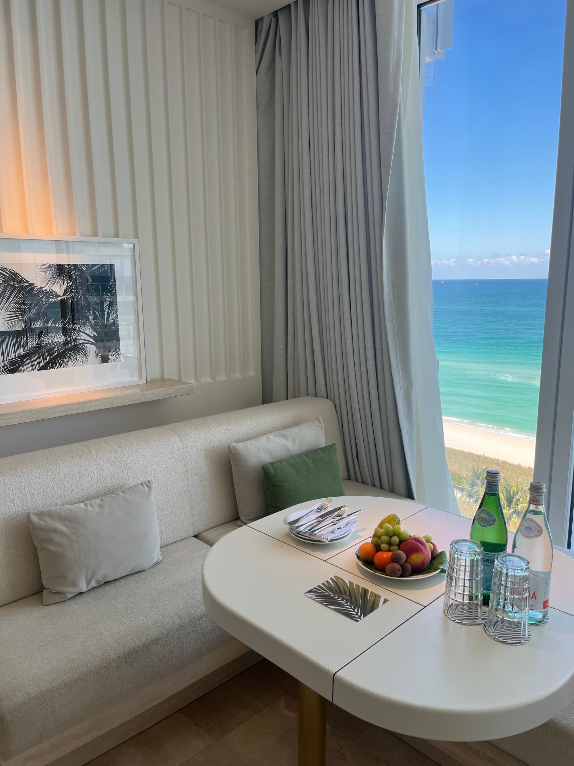 A hotel bedroom featuring white paneled walls, decorative artwork, a white couch with three decorative pillows, a table with snacks and beverages and window with curtains overlooking the turquoise blue water.