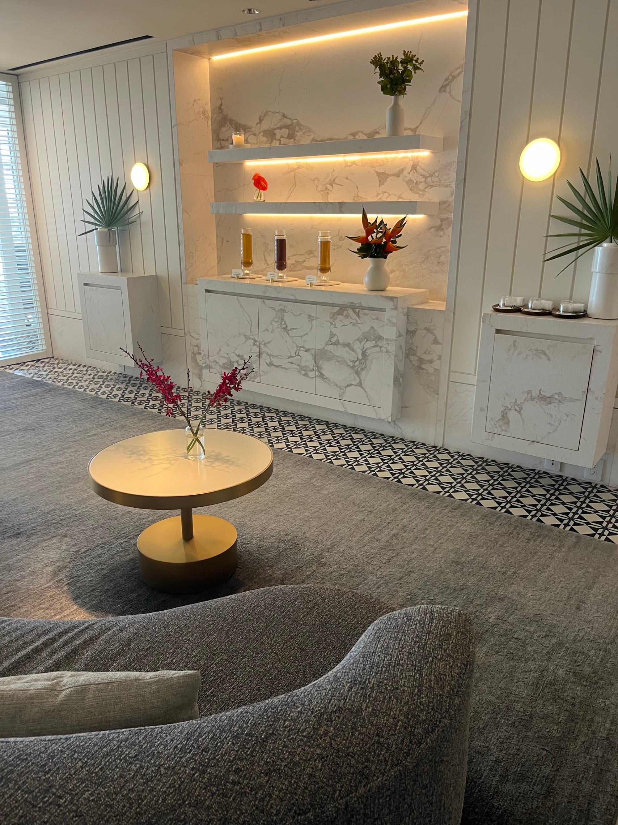A hotel spa with tiled flooring, a rug, a gold coffee table with a red decorative plant on top of it, grey couch, sconces, palm leaves in vases, marble detailing on the walls and lit shelving.