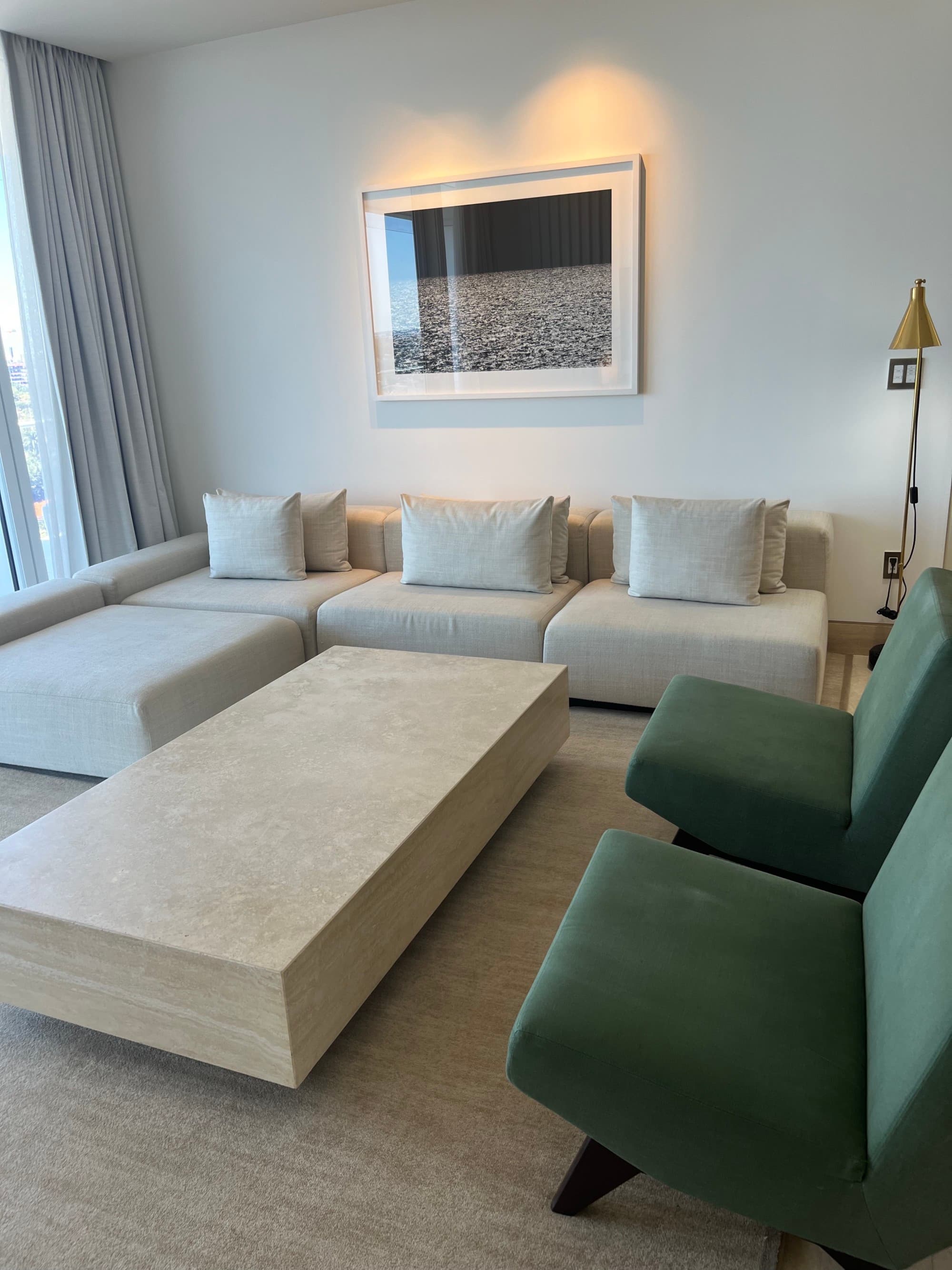 A lounge area in a hotel suite with a white couch, two green lounge chairs, artwork hanging on the white wall, a floor lamp in the right hand corner and a rectangular coffee table in the middle of the room.