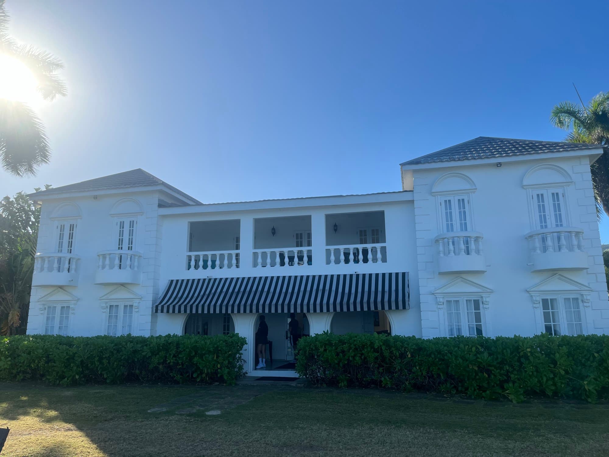 The exterior of the hotel, a simple white building with balconies, a blue awning and a green lawn.