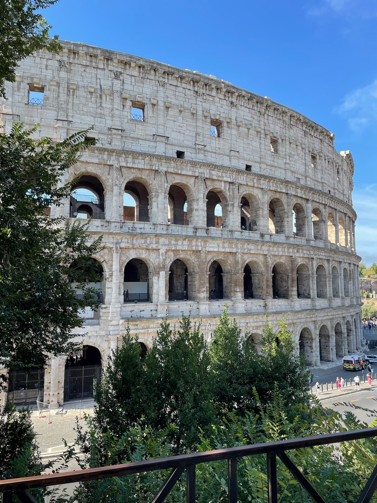The Roman Colosseum from a balcony across the street with trees in front.