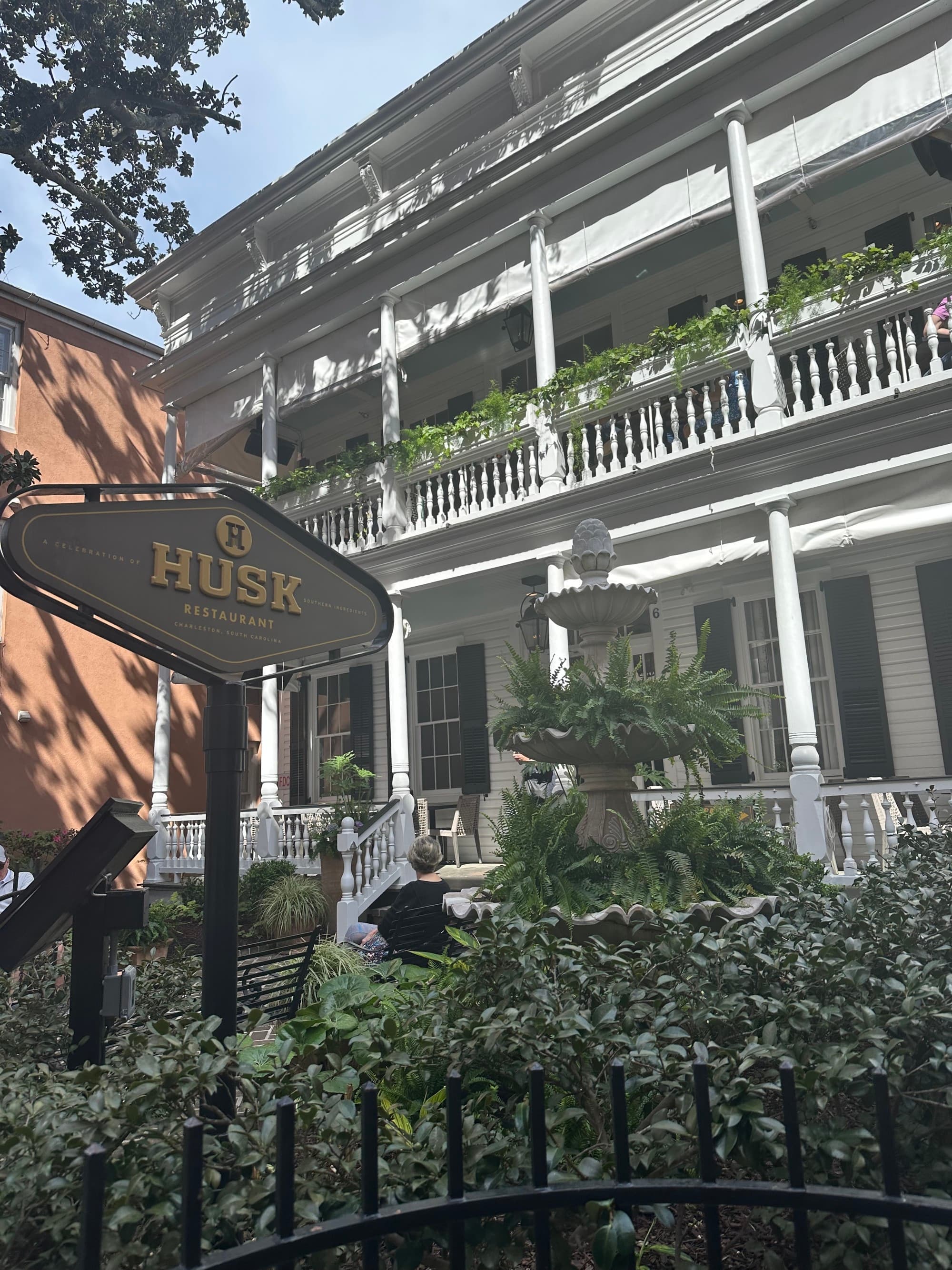 A restaurant exterior with white pillars, balconies, plants, bushes and a sign that says "Husk."