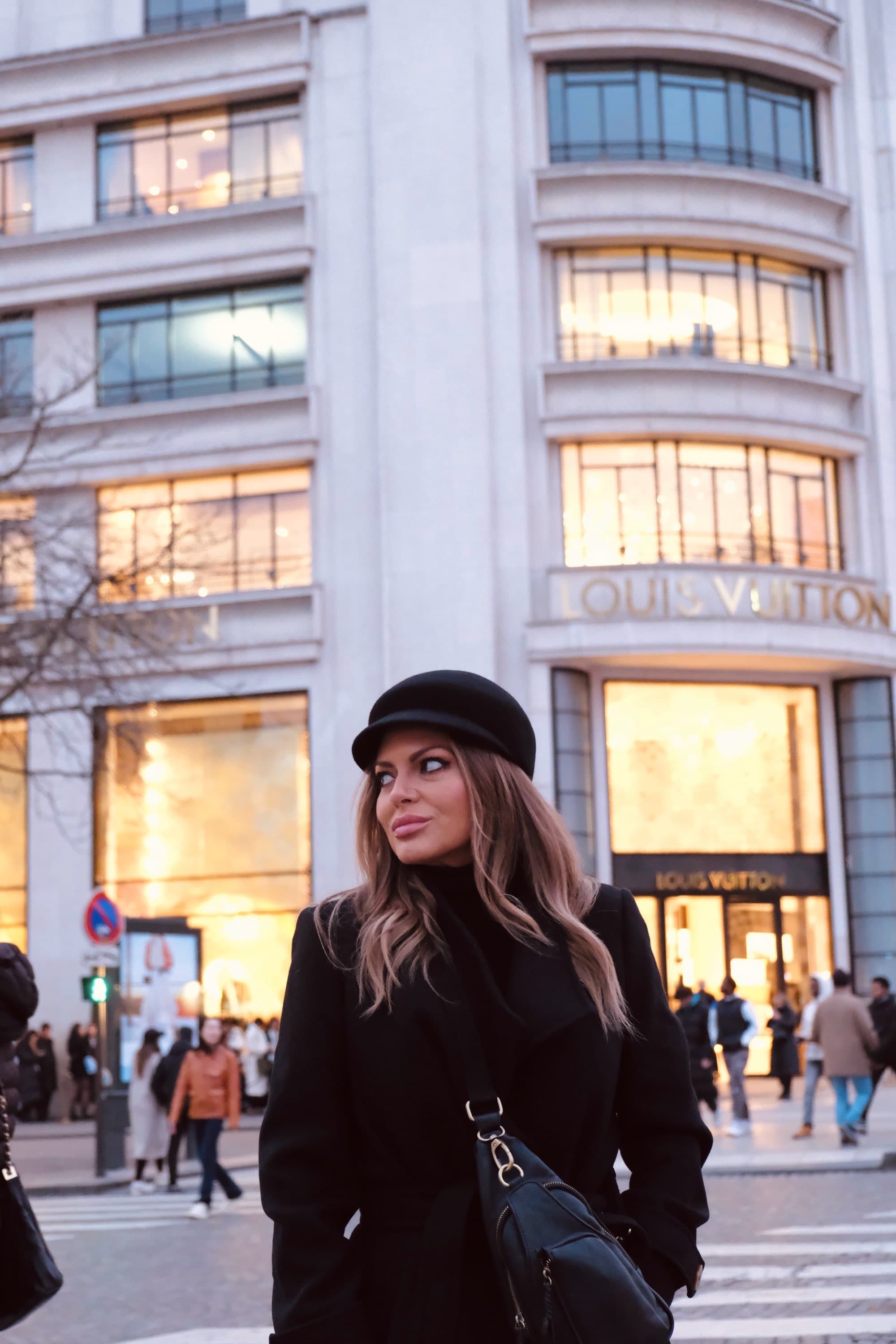 Travel advisor Kris in a black coat and hat posing in a crosswalk in front of Louis Vuitton in one of the best shopping areas in Paris.