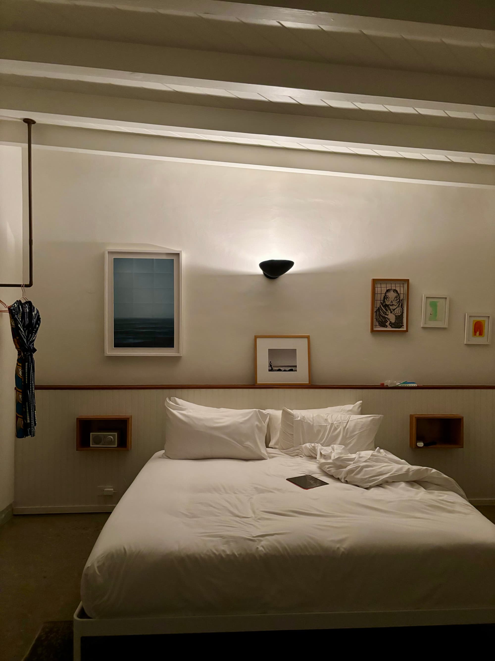 A white bed in a hotel bedroom with art on the walls and a light shining on the wall above the bed.