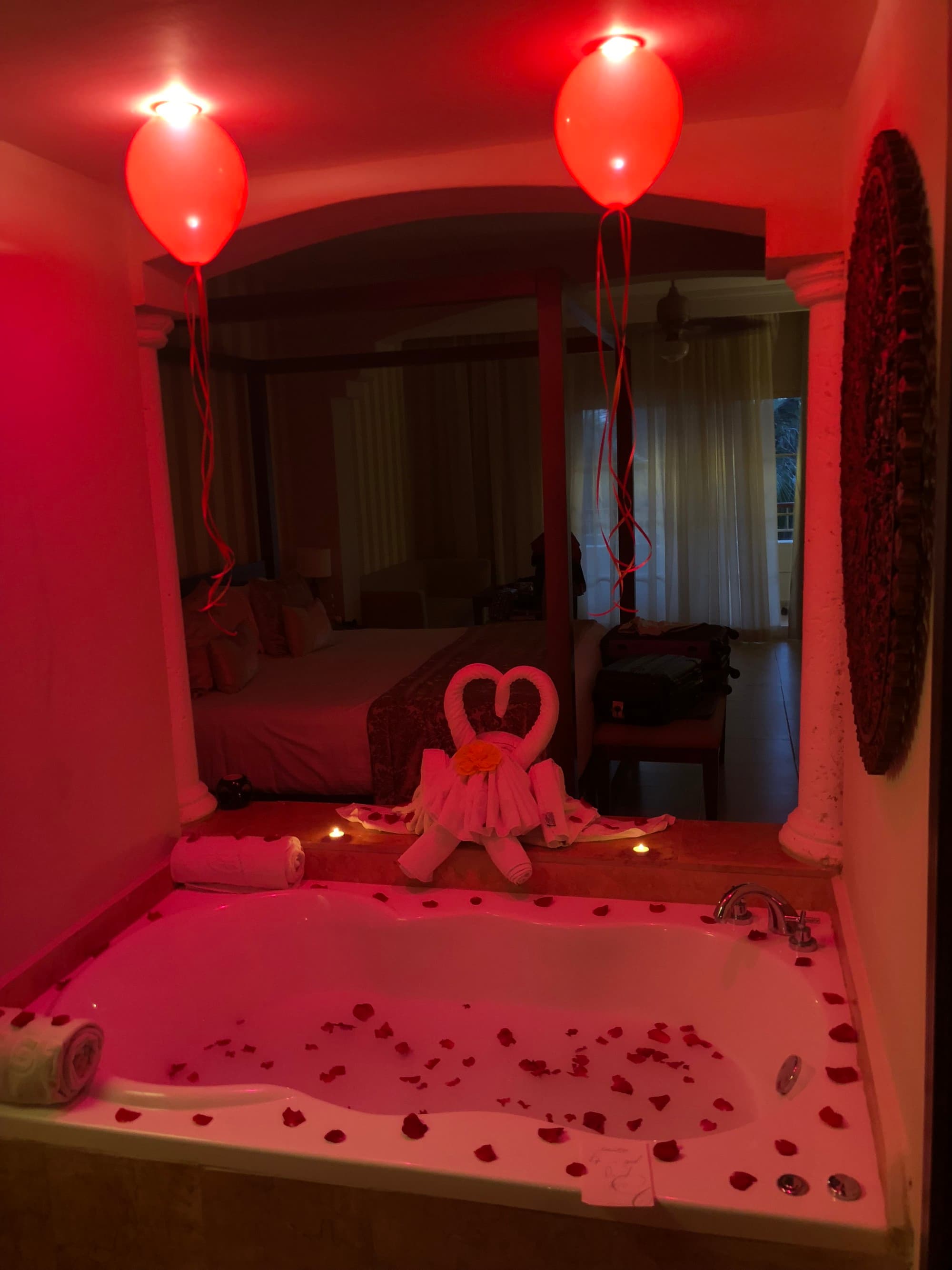 A bathtub with red balloons, red lighting, rose petals, heard shaped towels and a view of the bedroom in the background.