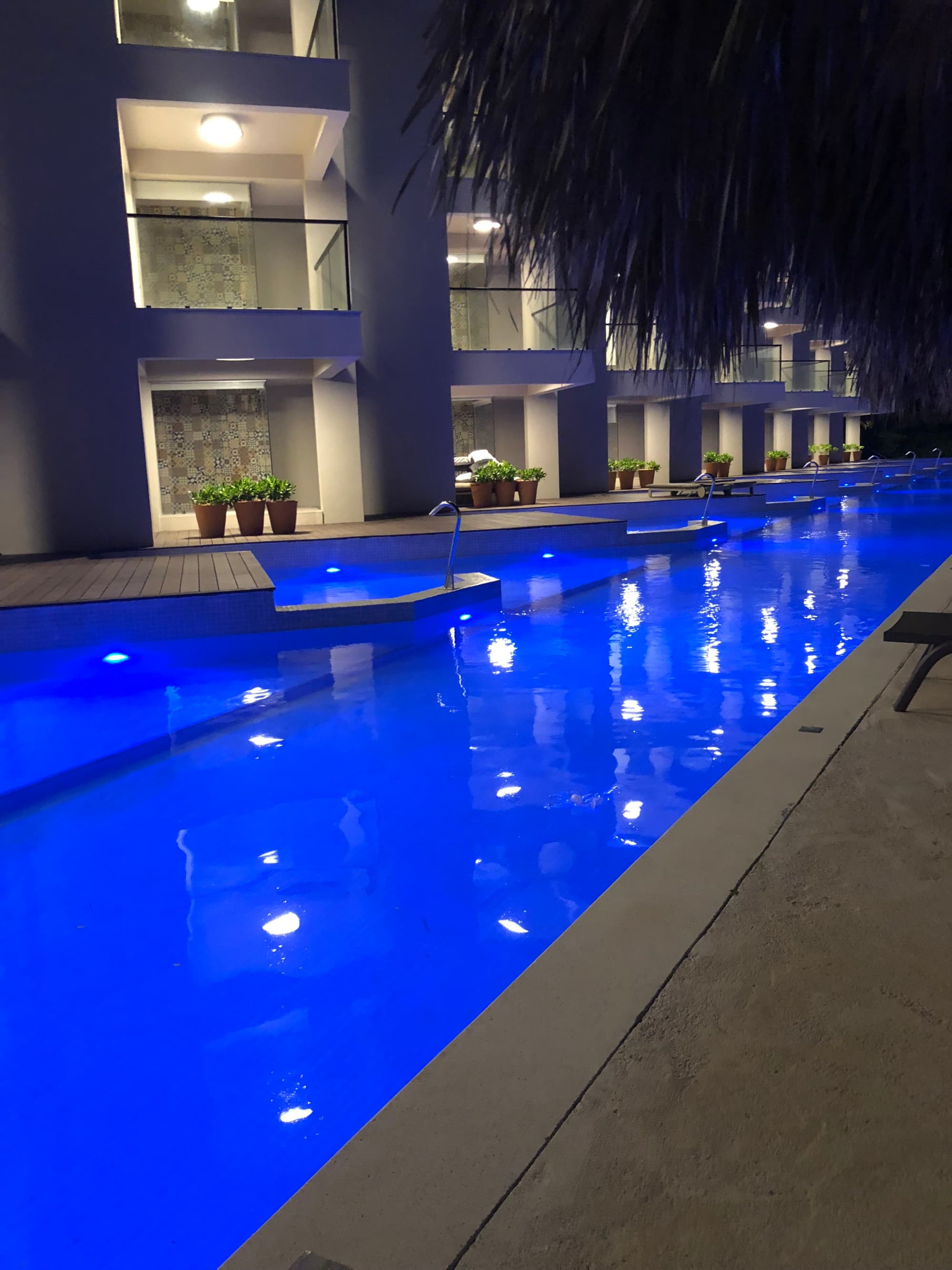 A swimming pool lit up with blue lights at nighttime. There are hotel balconies in the distance.