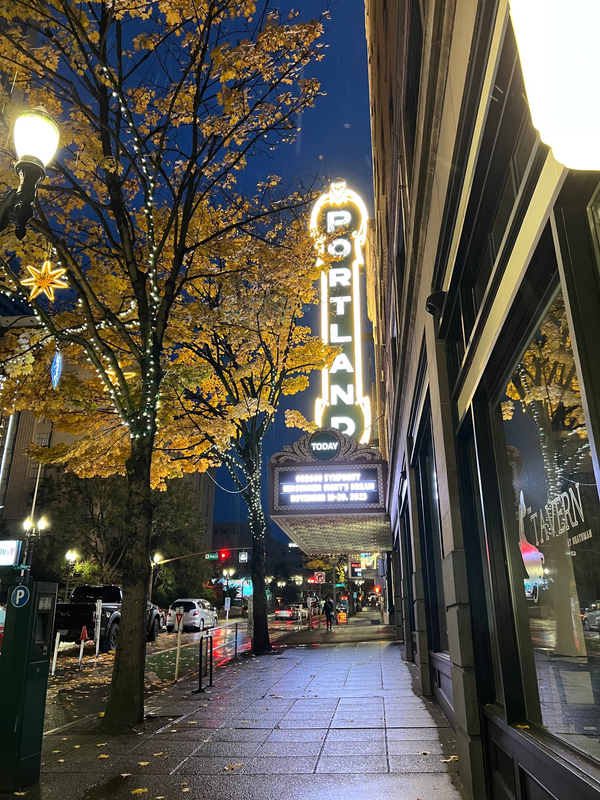 A view of a theatre sign that says Portland it lit up gold lettering next to a city sidewalk and trees.