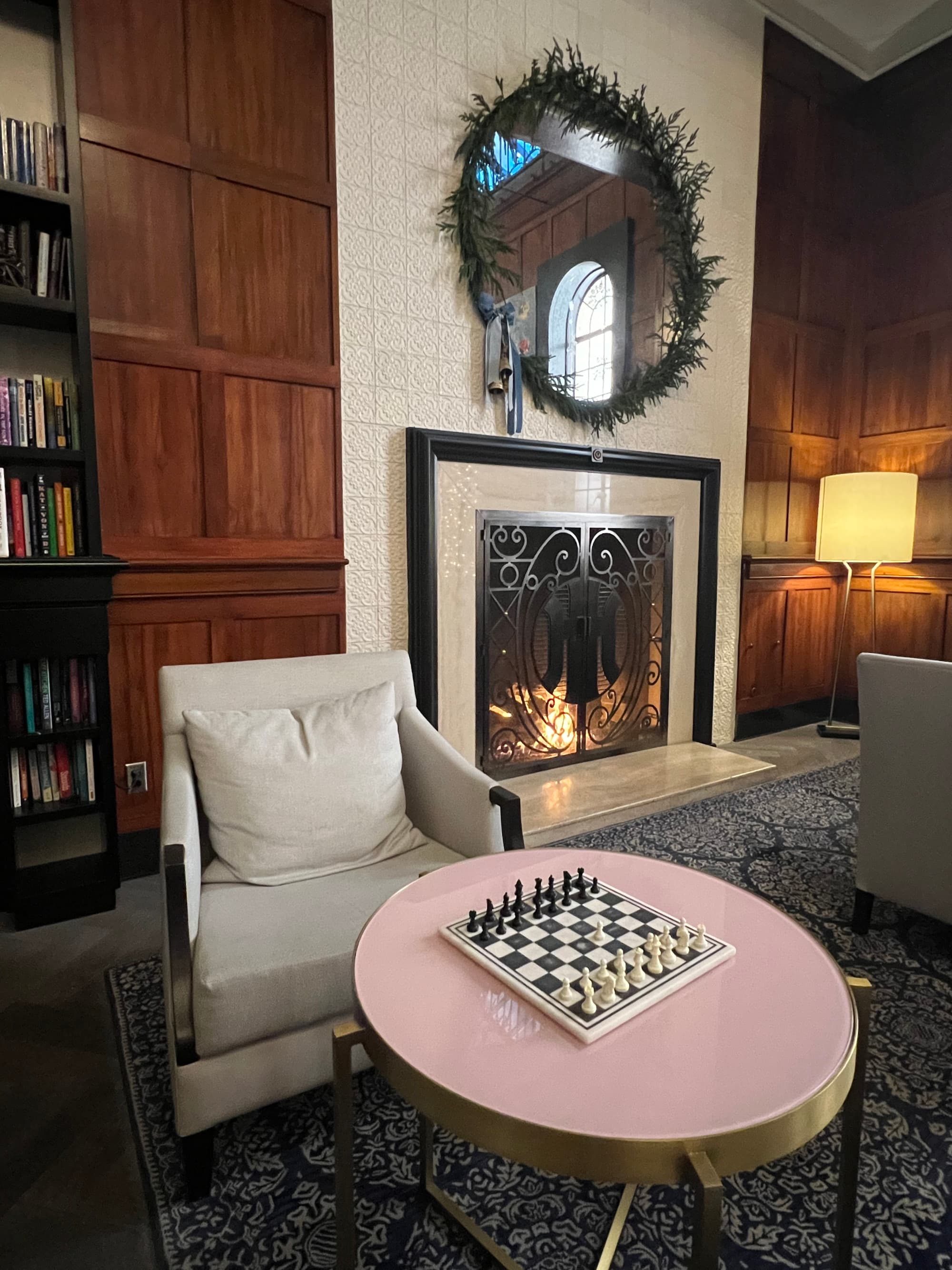A view of a hotel lobby with a fireplace, round mirror, wood paneled walls, white chair, round pink table and chess game on top of it.
