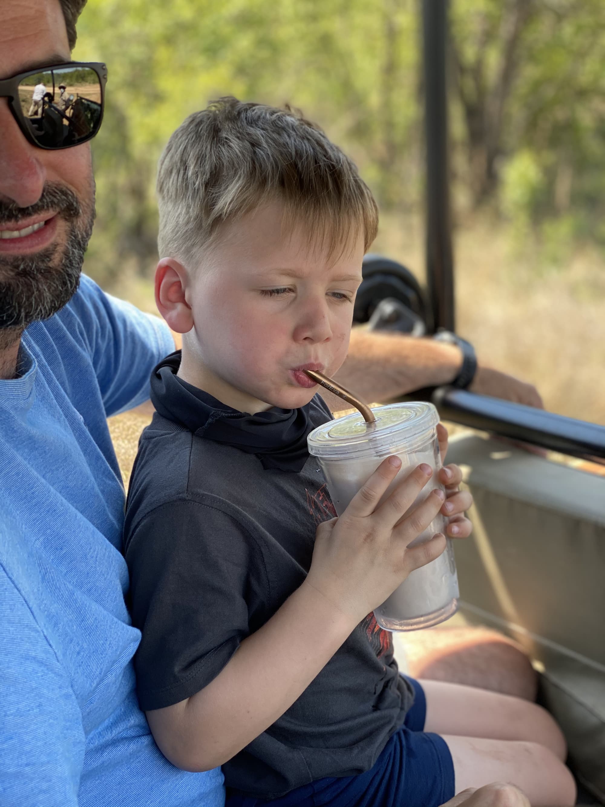 Child in a safari vehicle on his father's lap while drinking a milkshake