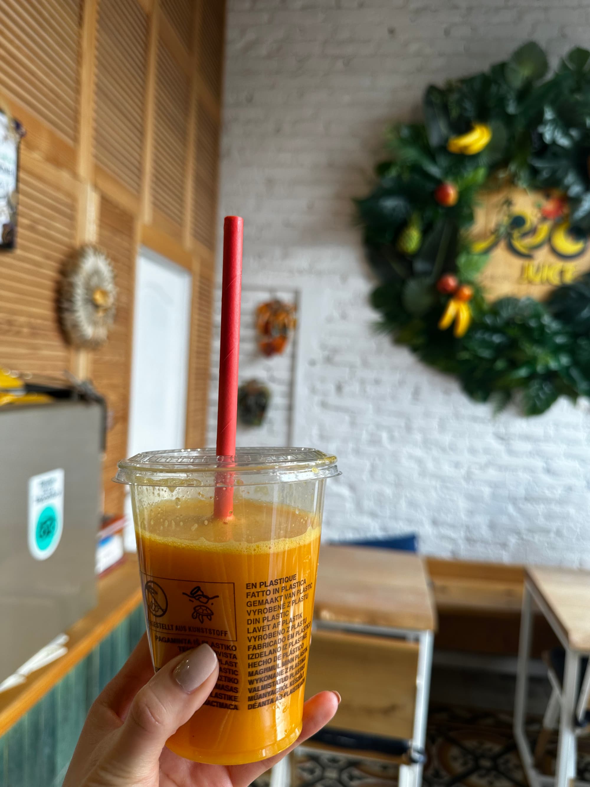 Someone holding up an orange juice with a red straw. There is a cafe with a white brick wall, wreath and wooden tables and chairs in the background.