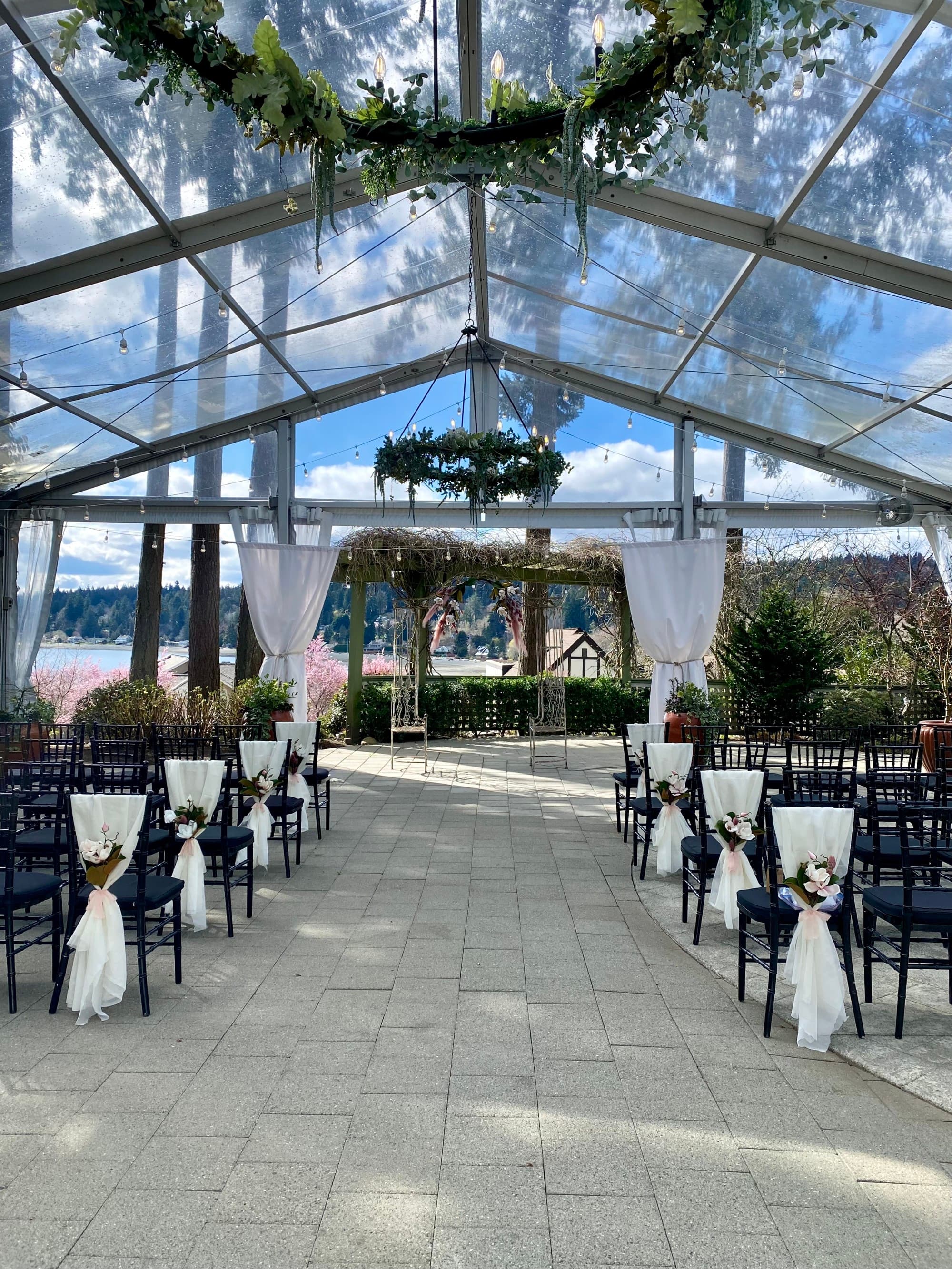 A wedding venue with white cloth chairs, tiled flooring, woven wreaths hanging from a glass ceiling and trees in the background.