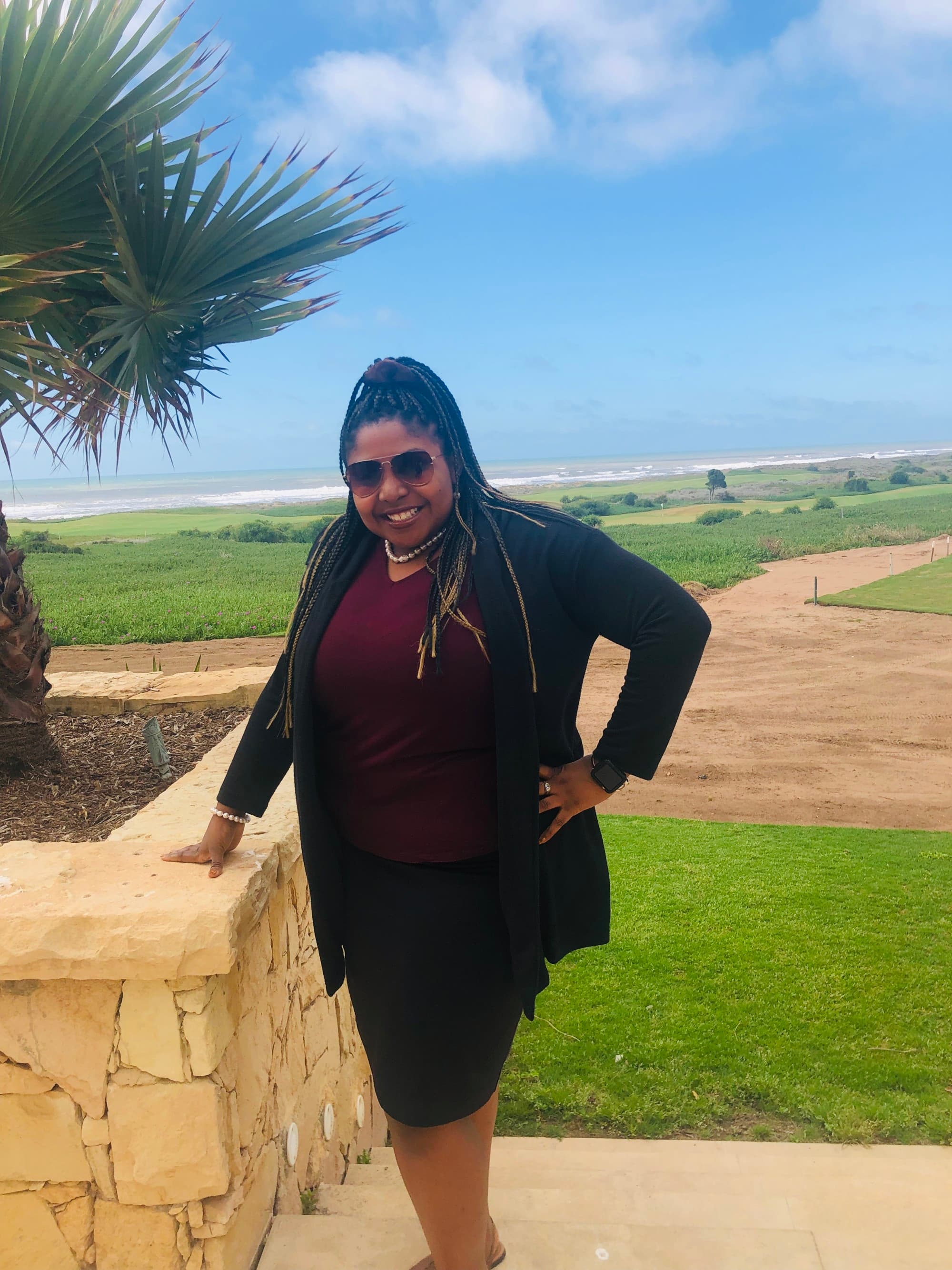 Tysheda in a black skirt-suit, posing by a stone wall, with a dirt road going through a green field in the background.