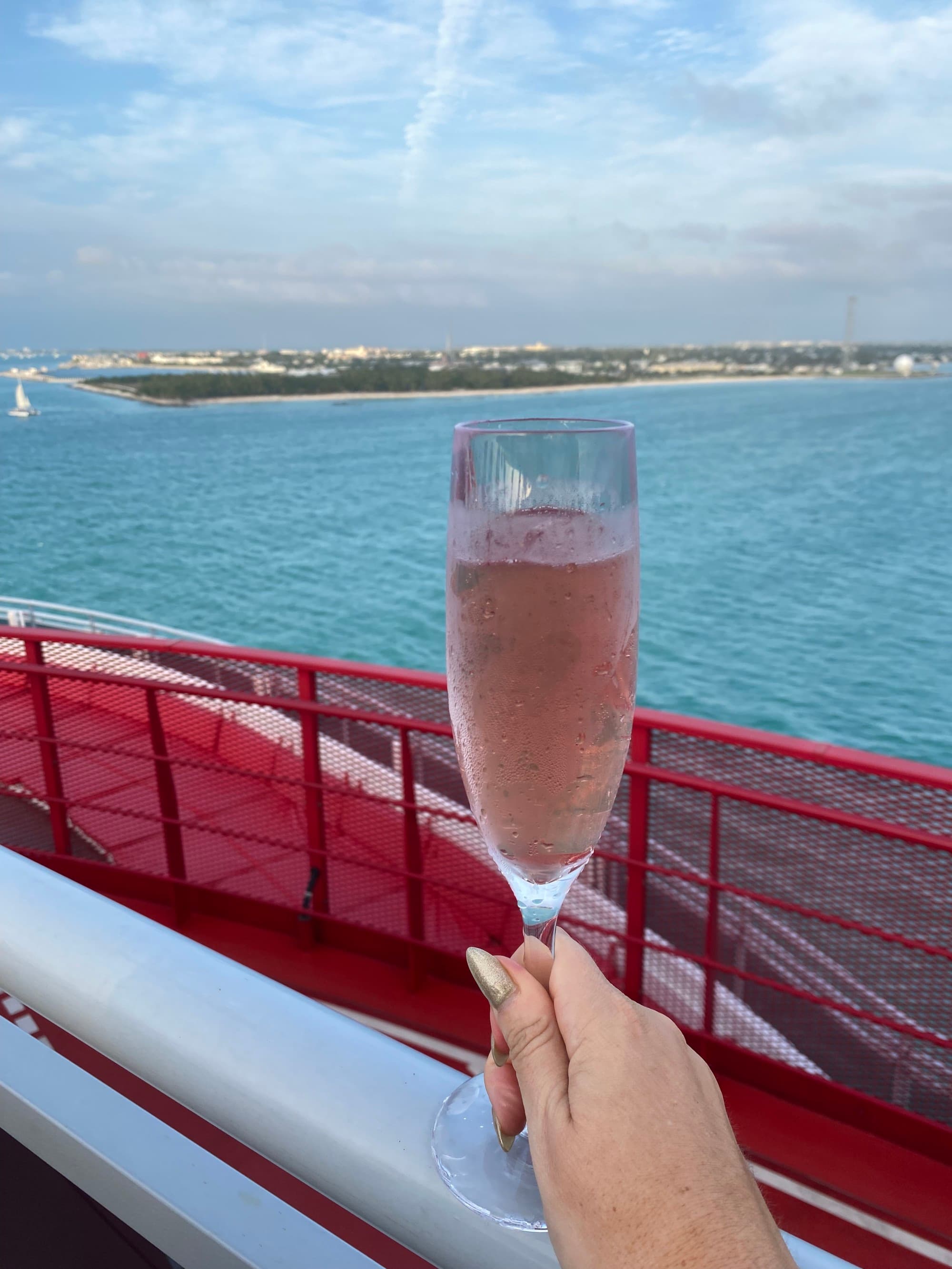 A picture of a hand holding a champagne flute with a light pink liquid inside of it. There is a red railing and view of the blue sea in the background.