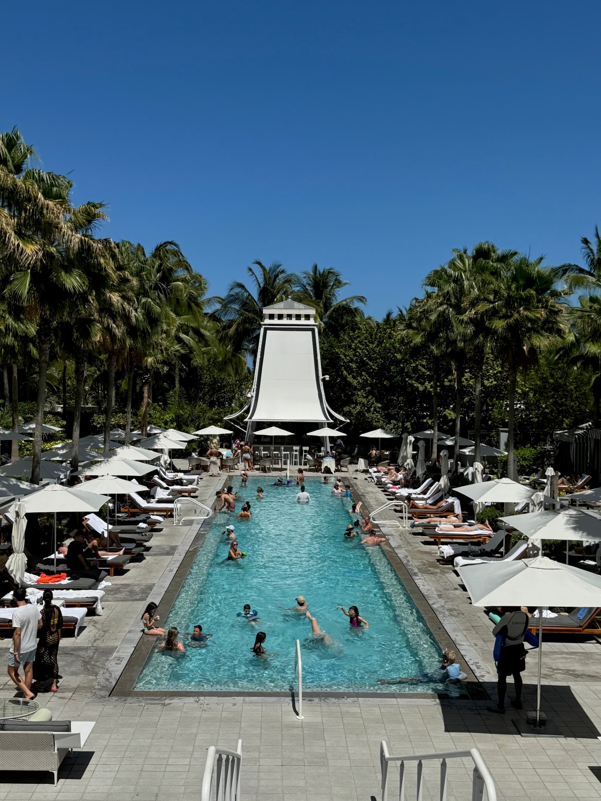 An image of a swimming pool with a large slide in the distance surrounded by lush palm trees. There are cabanas, people swimming and lounge chairs in the surrounding area.