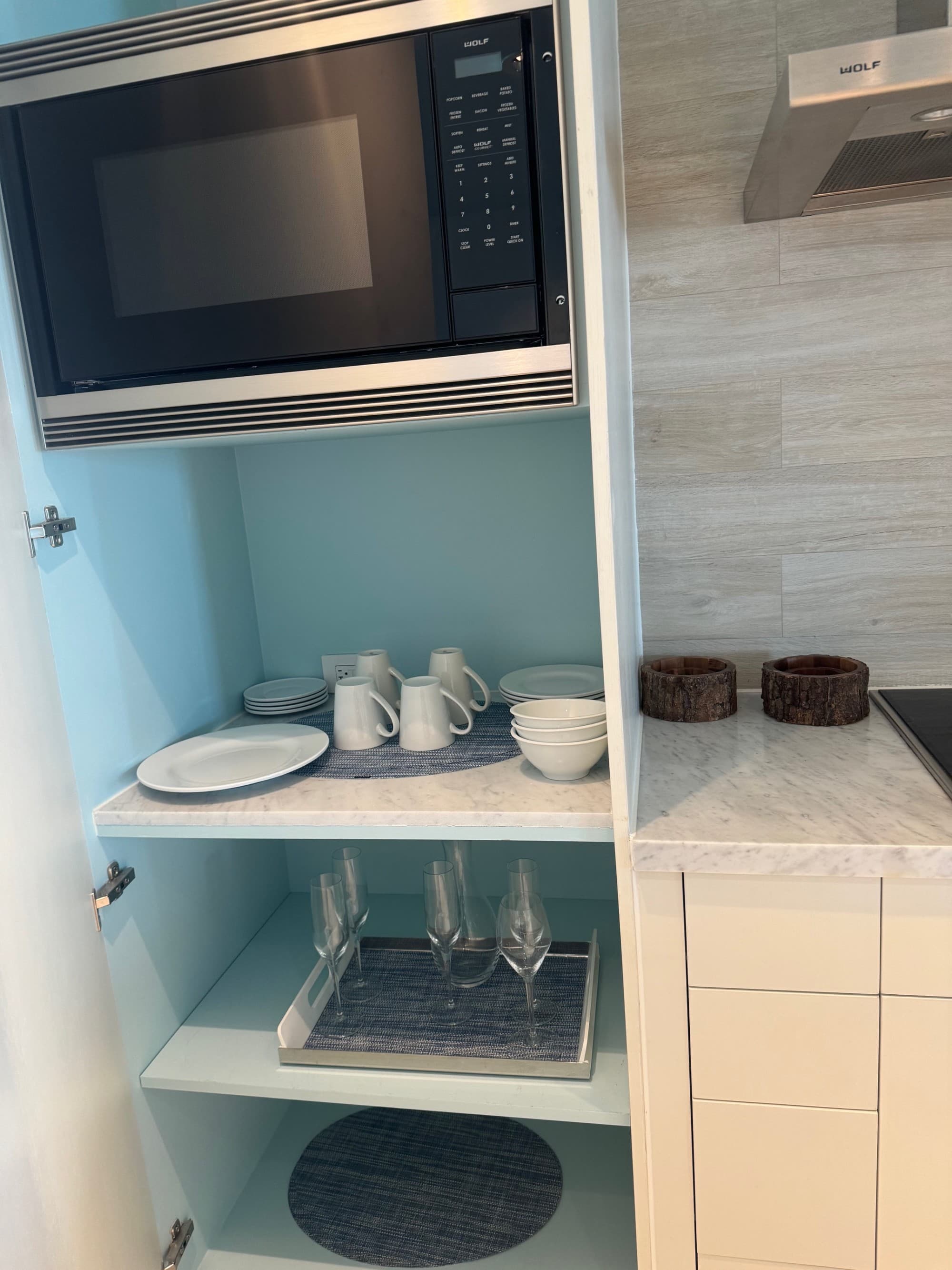 An image of a kitchenette with a black microwave, light stone work and cups, plates and bowls inside of a cupboard.