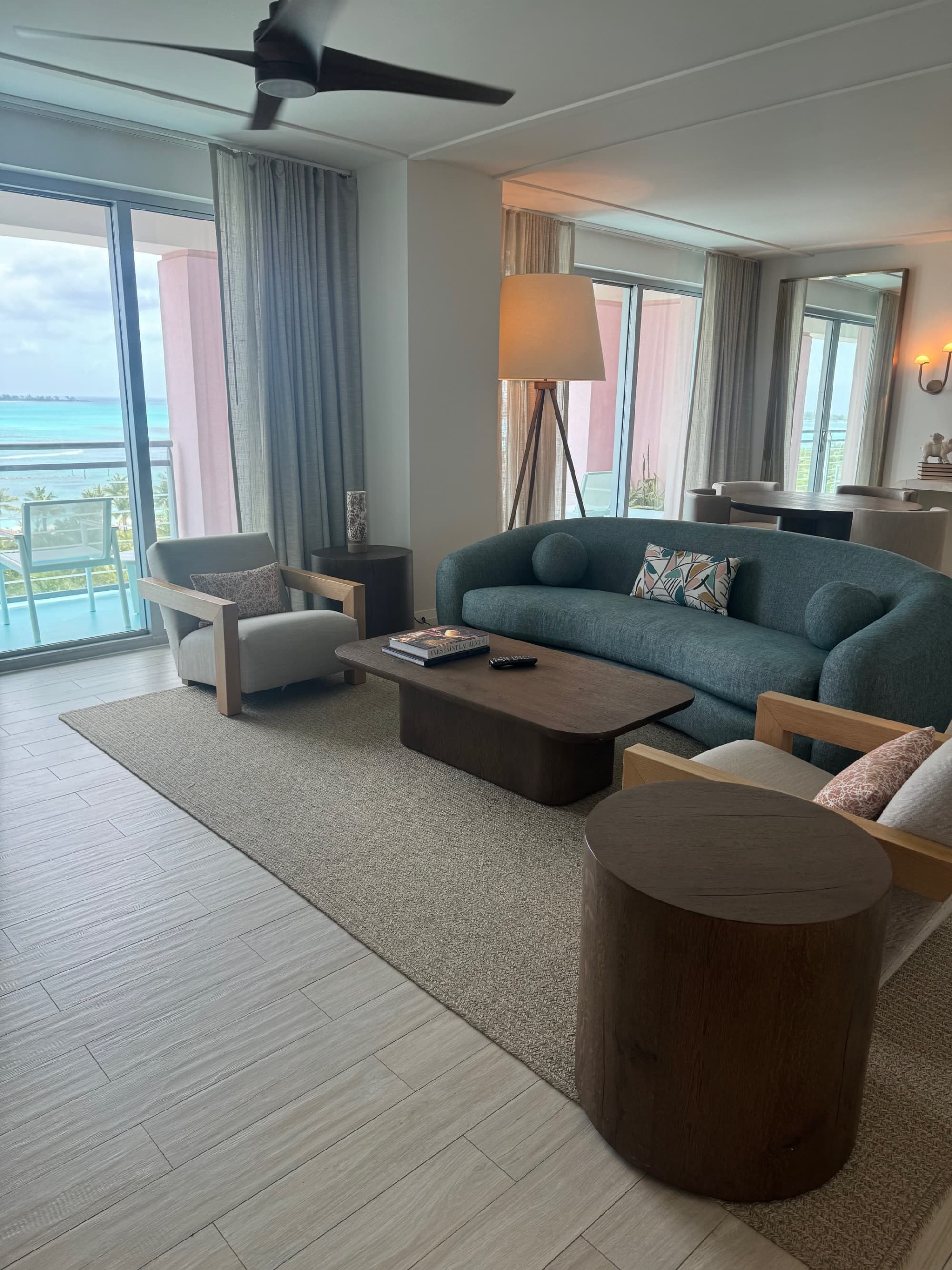 An image of a hotel suite with a blue couch, wooden coffee table, round side table, lounge chairs, a rug, ceiling fan, floor lamp and a window looking out to a view of the sea.