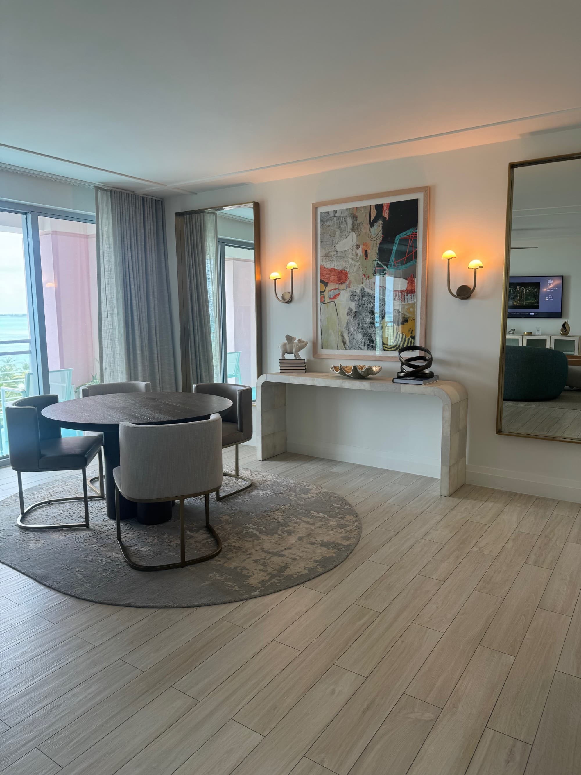 A hotel suite with a round table surrounded by four chairs, a rug, a console table, wall sconces turned on, artwork on the wall, mirror and window with curtains toward the left side of the image.