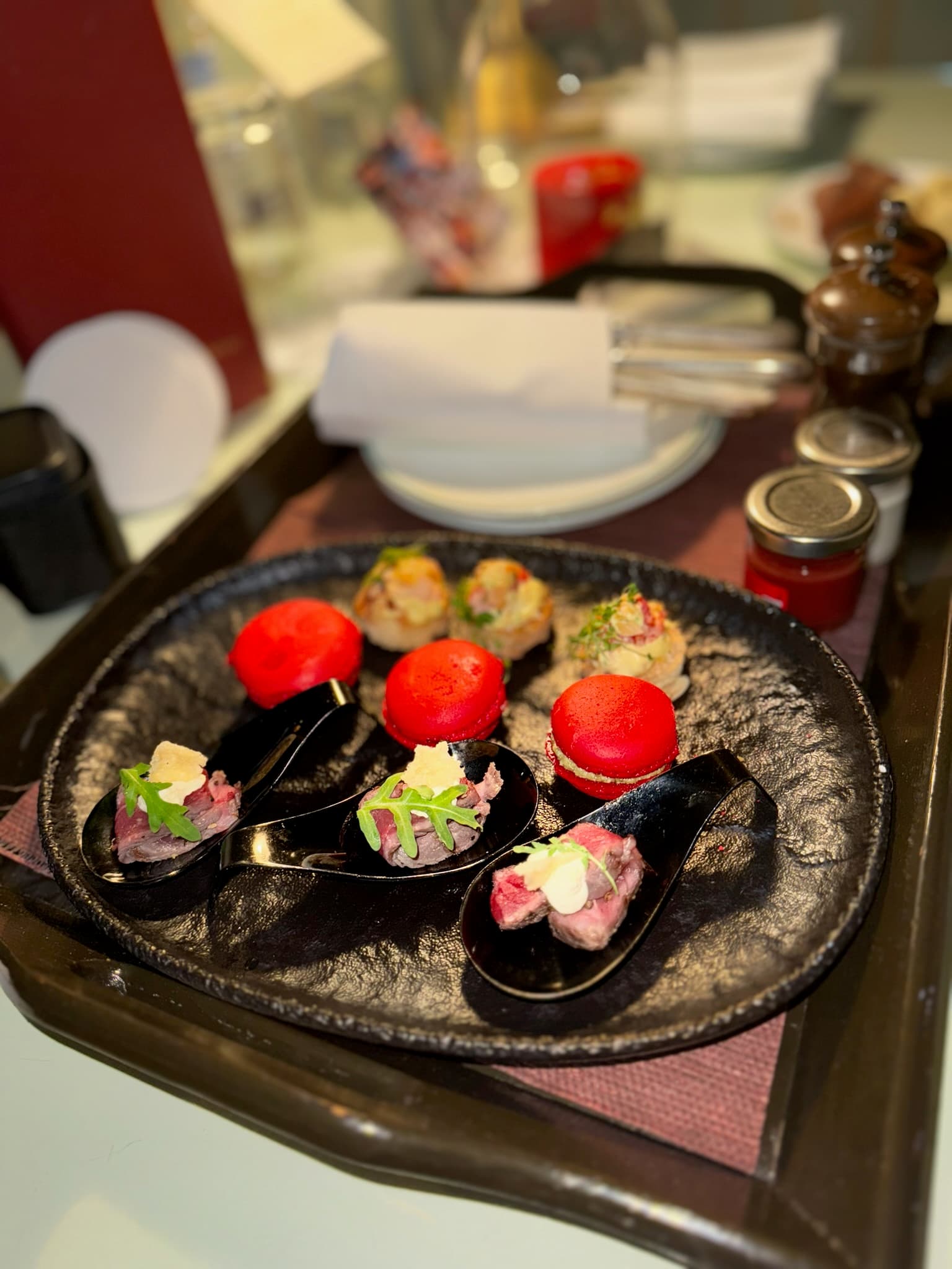 A plate of spoons holding rolled meat, topped with green herbs, with red macaroons behind the spoons, and another bite-sized appetizer behind the macaroons.
