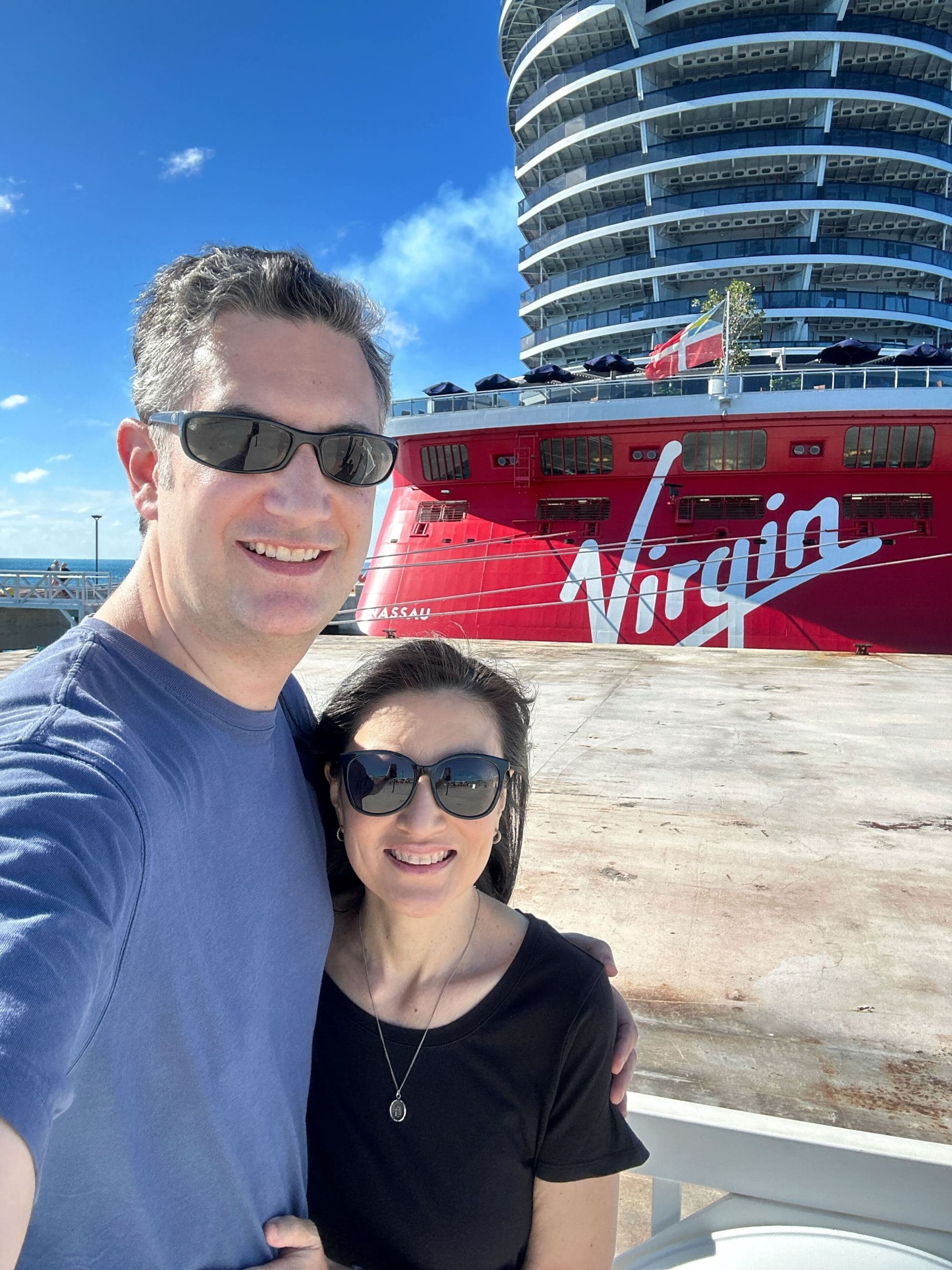 a man and a woman wearing sunglasses stand in front of a red Virgin Voyages cruise ship