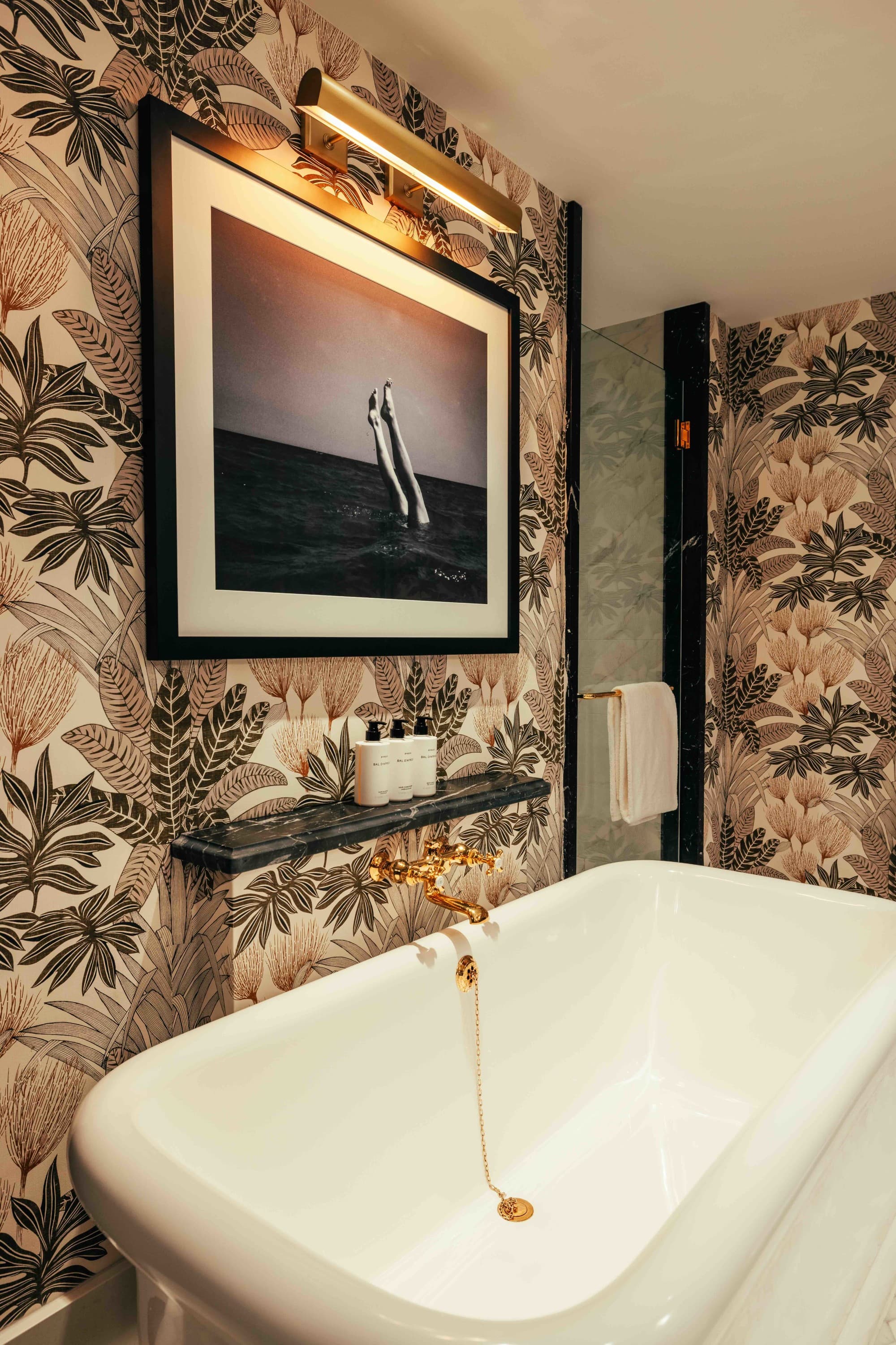 A white freestanding bathtub in a bathroom with tropical printed wallpaper and a black-and-white photo of a woman's legs sticking out of the sea.