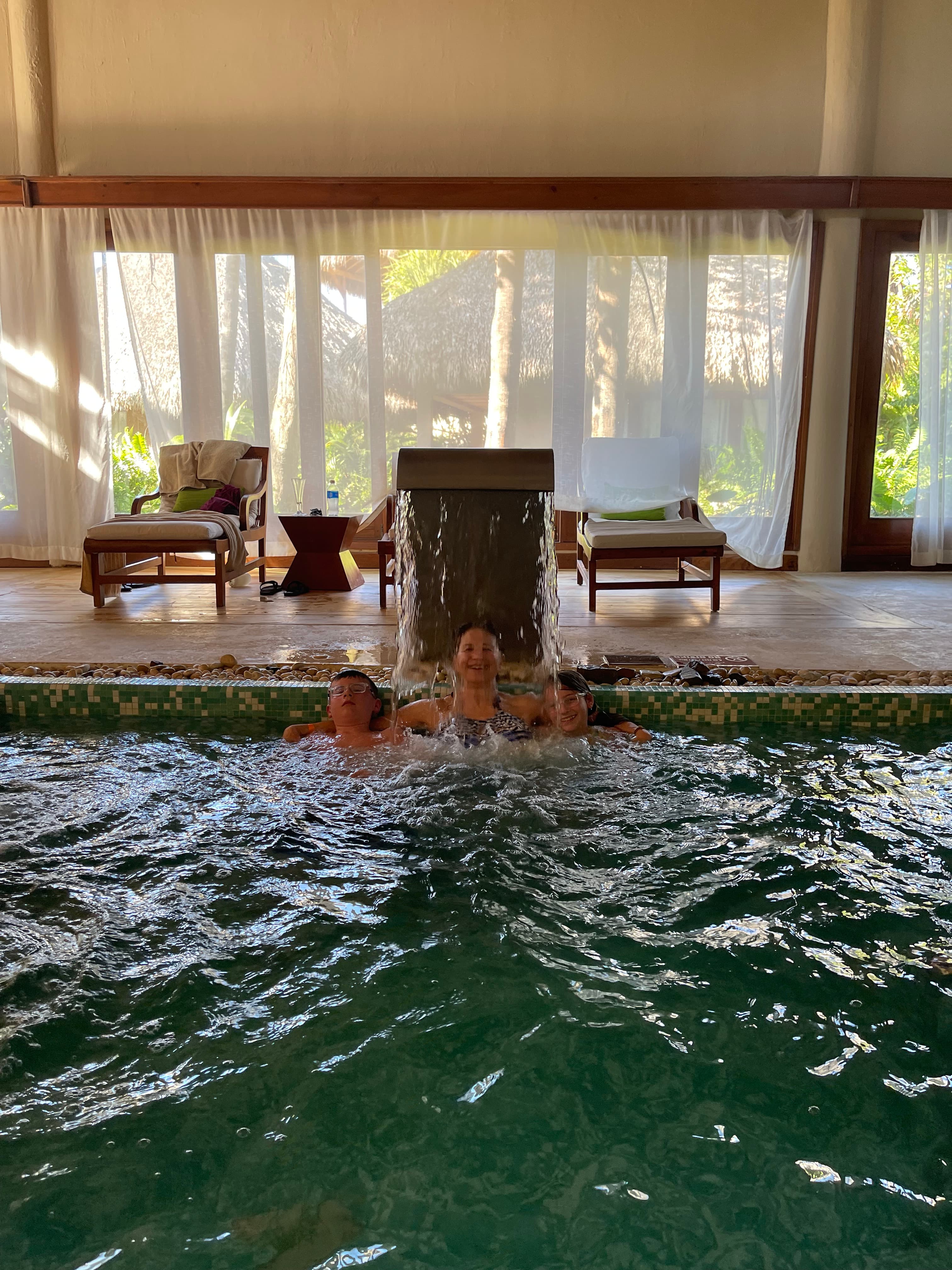 An indoor pool with a woman and two children standing under a small jet with sun loungers and large windows behind