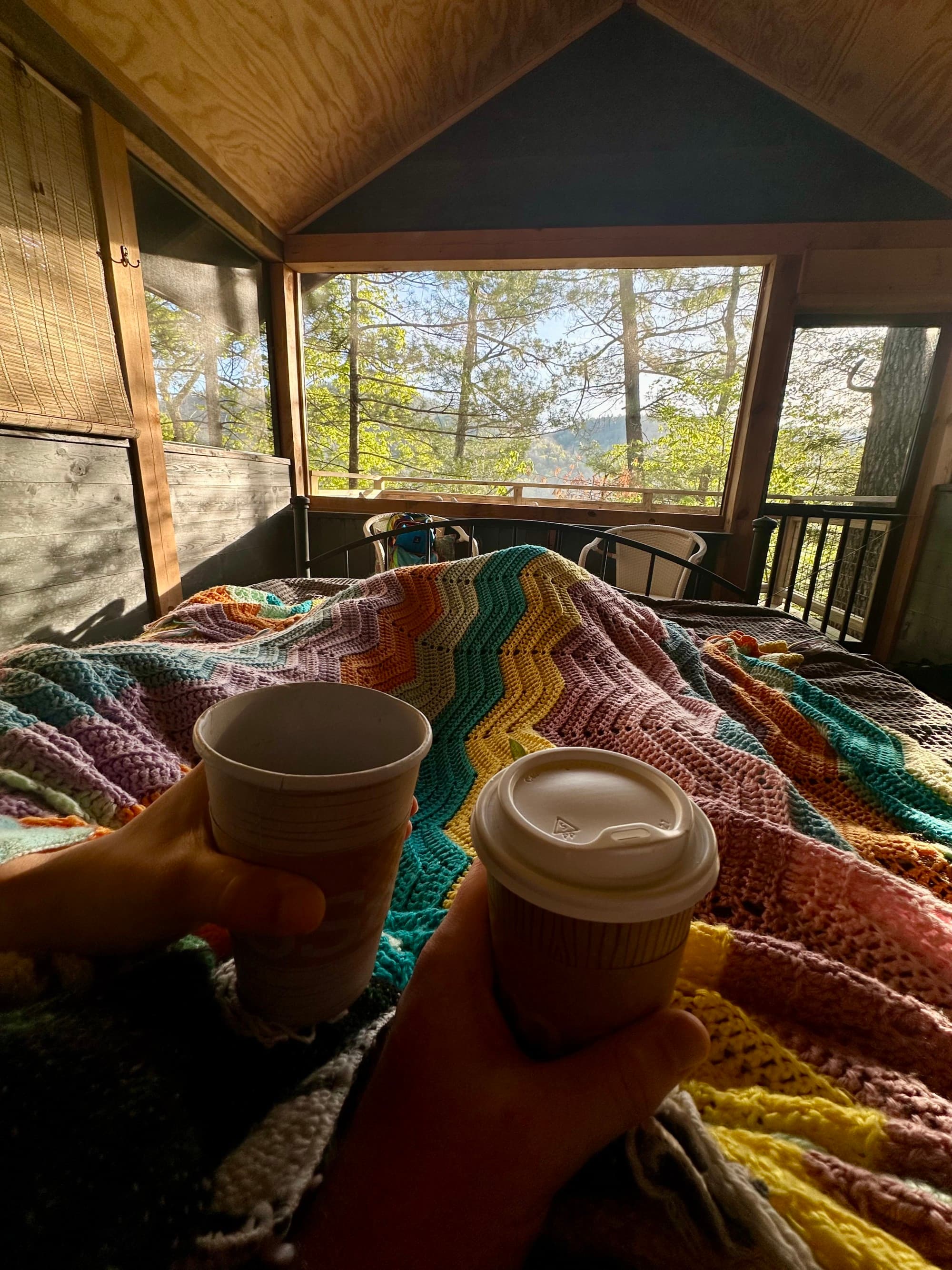 Two people holding disposable coffee cups in bed, with a multi-colored blanket, and forest view from the opposite window.