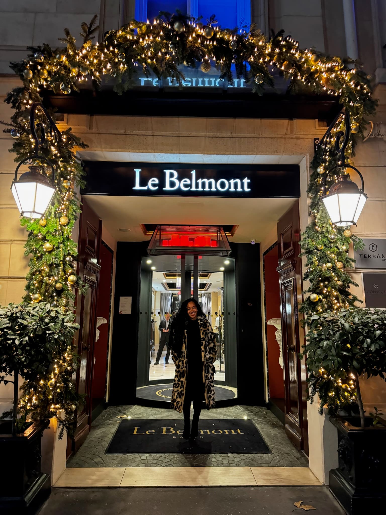 Tysheda in a leopard print coat standing outside the main door to Le Belmont, surrounded by lit-up greenery