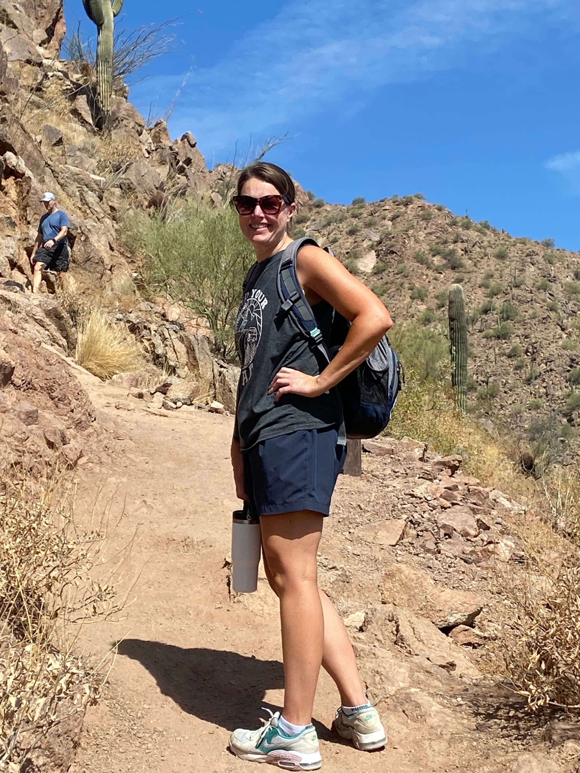 A woman in sunglasses, a tank, and shorts smiles on a mountain trail