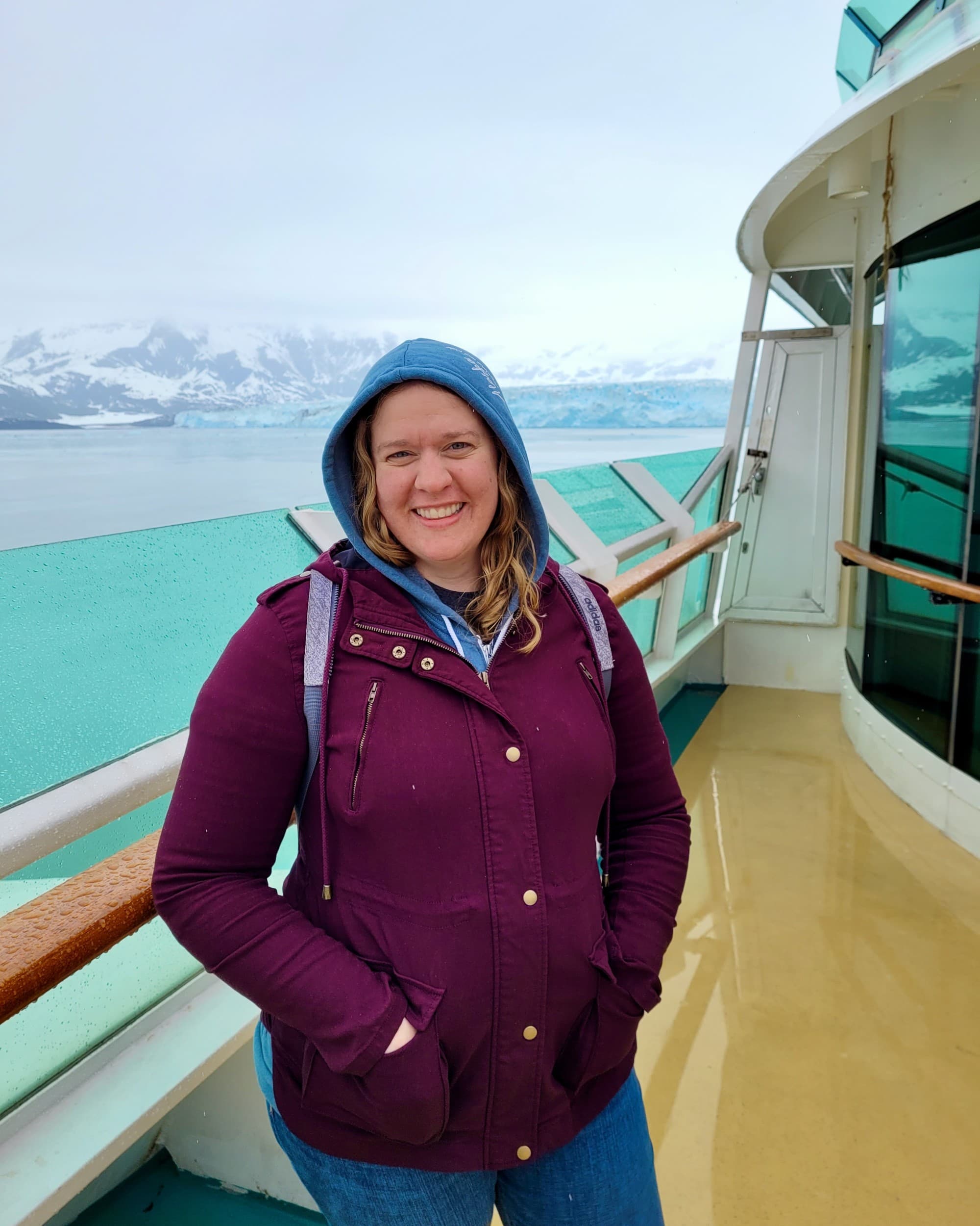 Advisor wears a purple rainshell on the deck of a ship with snowy mountains in the background.