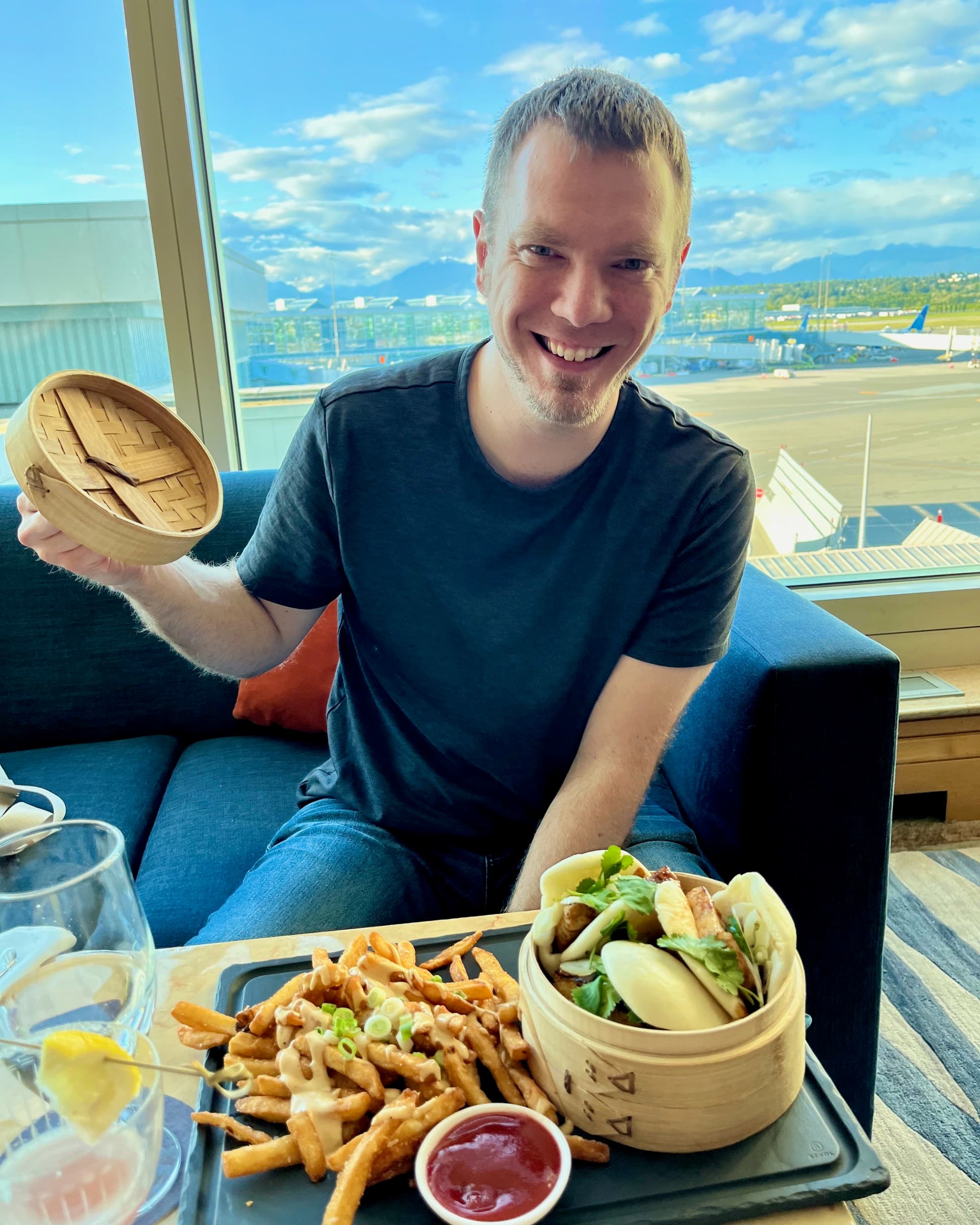 A man sitting with a plate of food in front of him sitting on a sofa with planes and a runway in the background.
