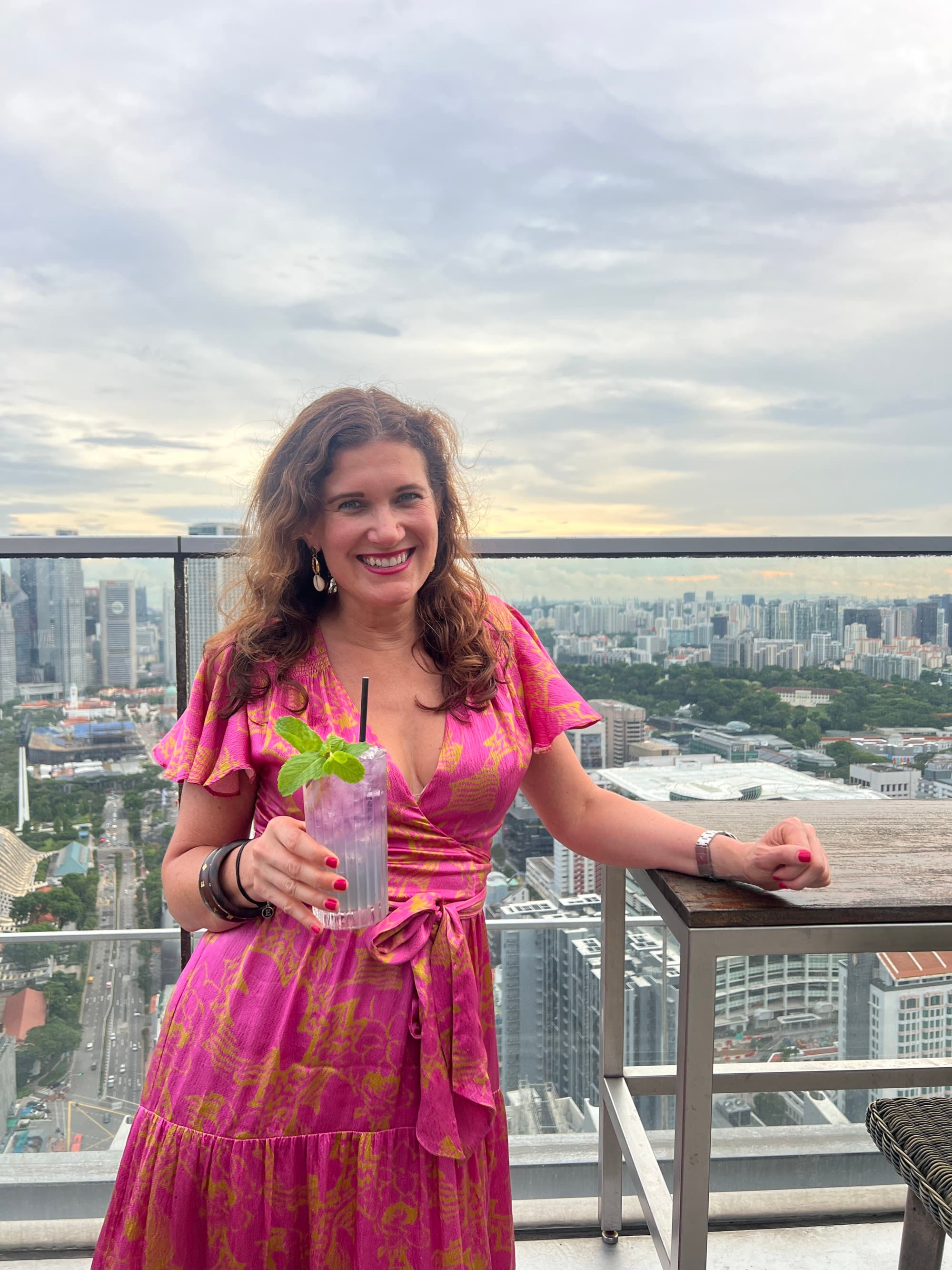 Advisor wearing a pink dress and holding a cocktail with a city skyline in the background.