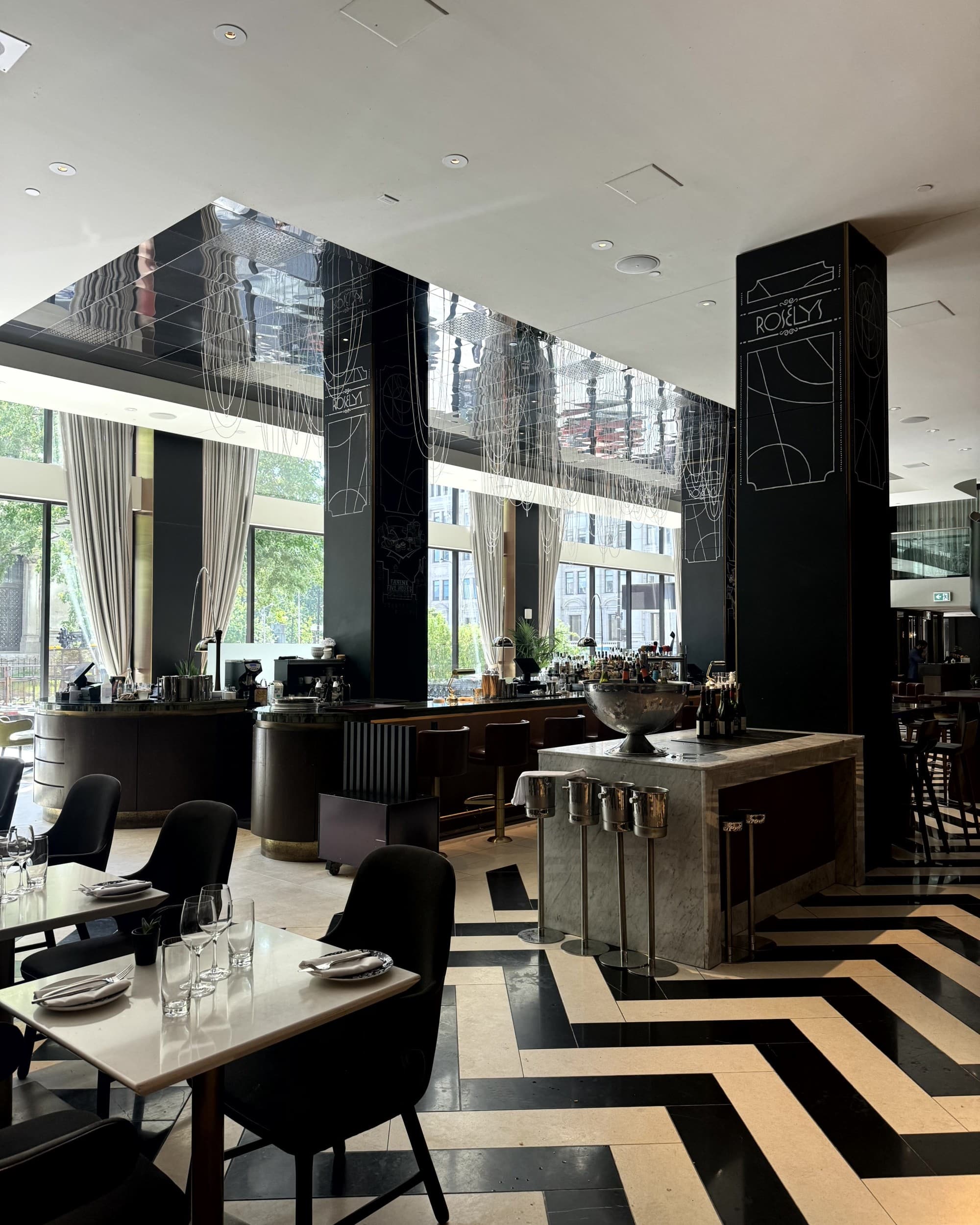 A view of a modern looking restaurant with black and white chevron tile, white tables and black chairs.
Large windows let in daylight at Gastronomic Delights - Roselys - Rondene Grinam