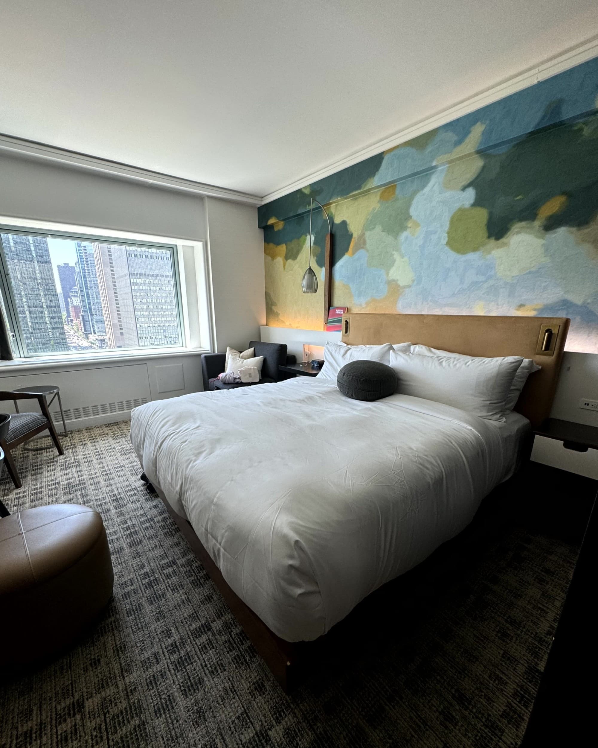 A view of a hotel room with a king size bed and a large green and blue mural on one wall.
The window looks out to the city. Accommodations - Room 2 - Rondene Grinam