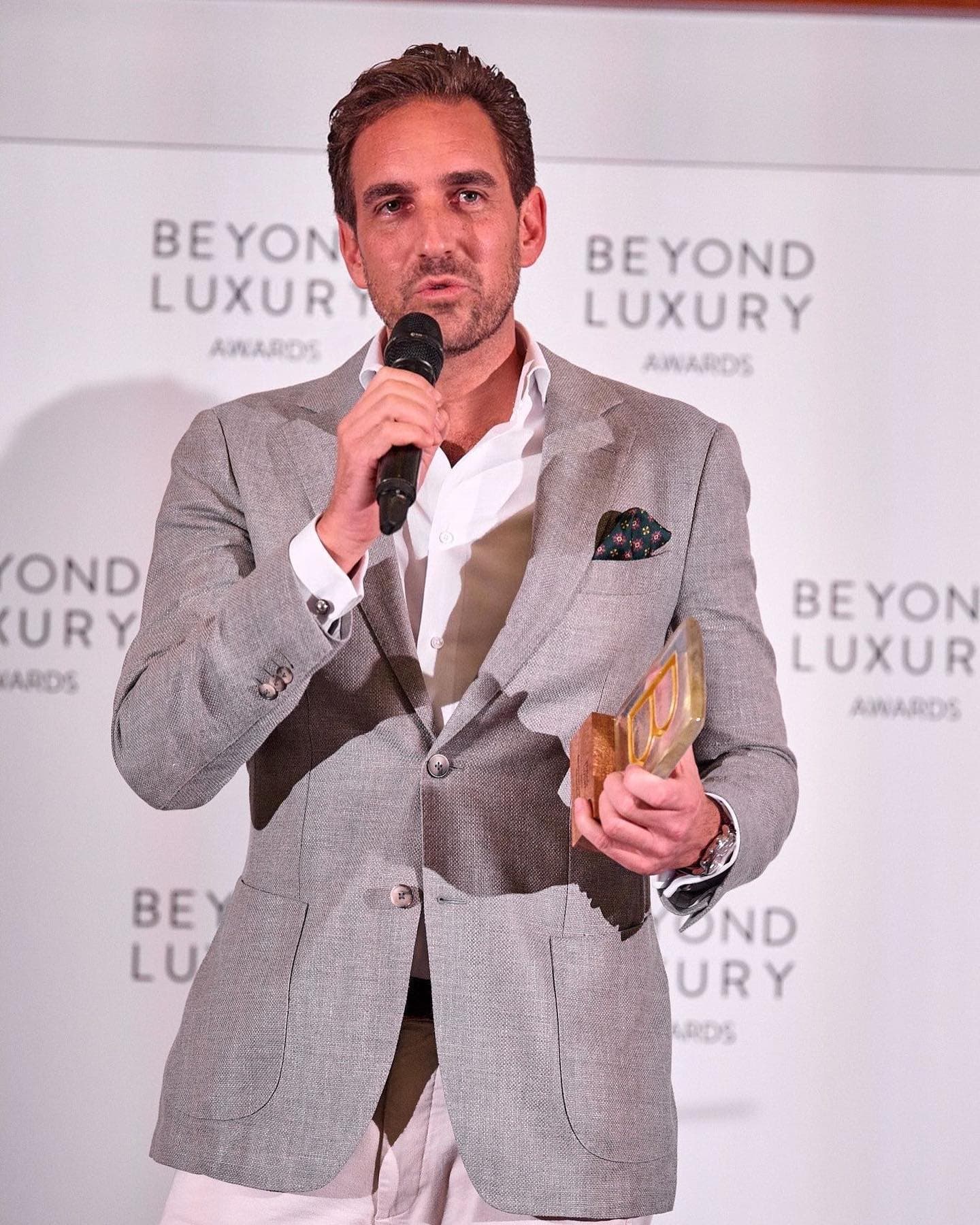 a man in a gray suit speaks into a microphone while holding a trophy