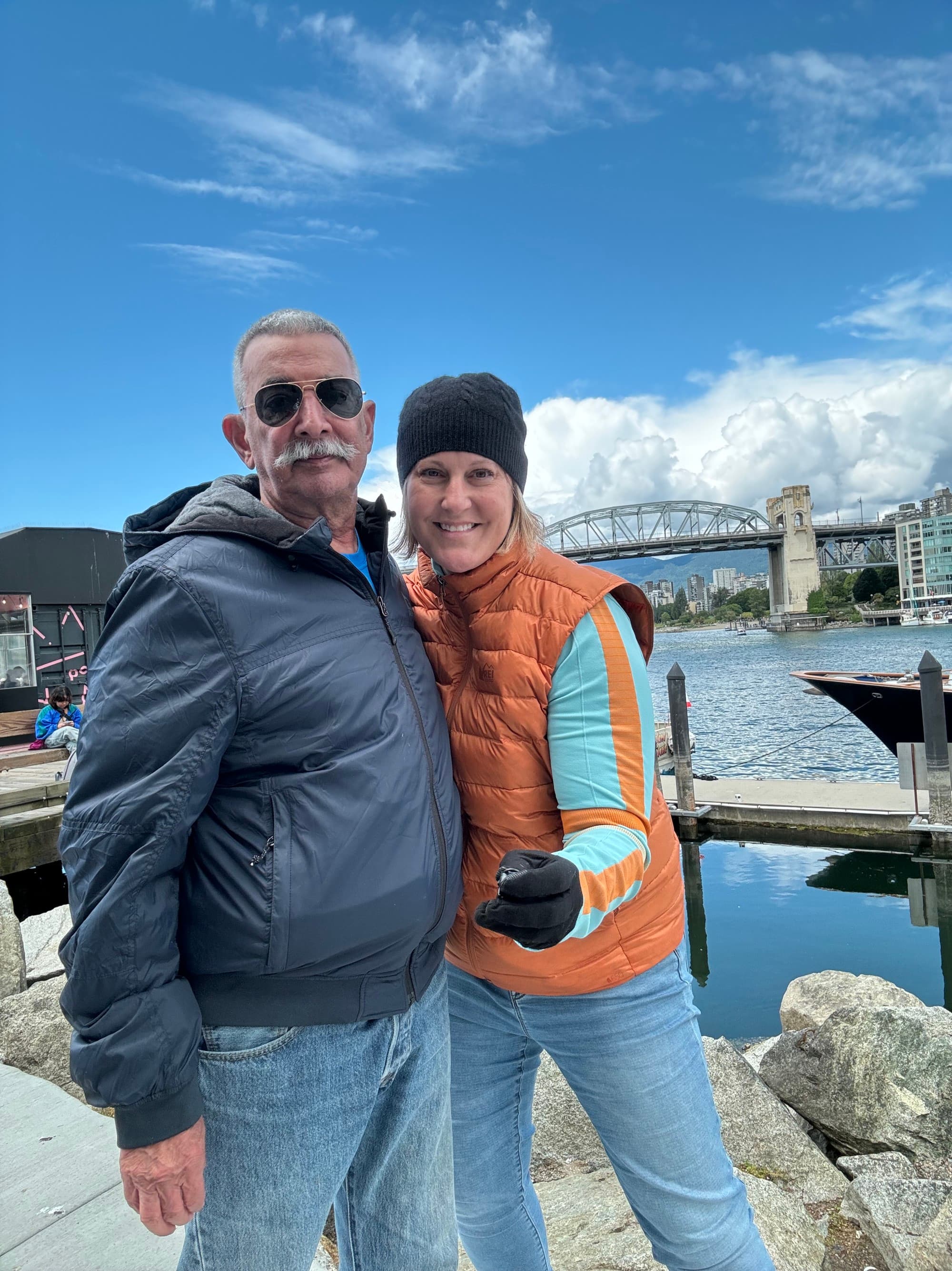 A couple wearing warm clothing posing in front of rocks, water and a bridge in the distance.
