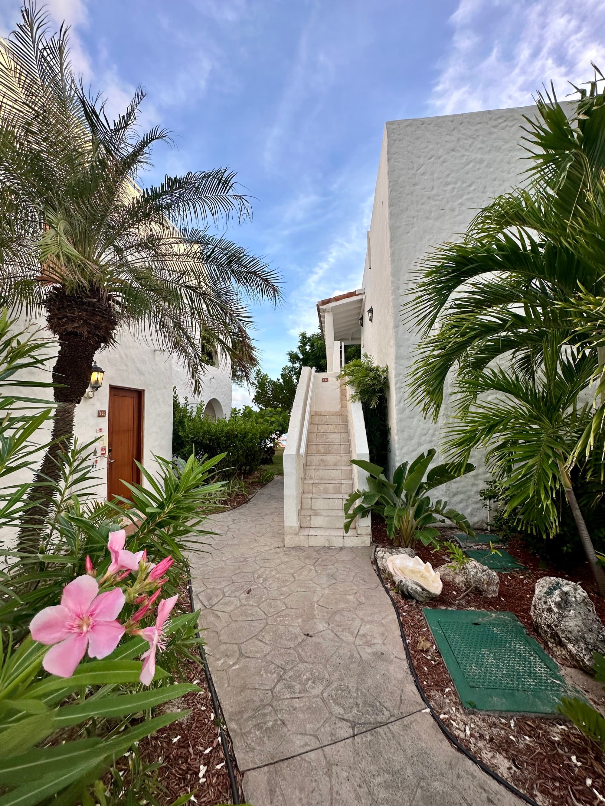 Outdoor walkway stretches along separate bungalows that are lined with local flora and palm trees on a clear day dotted with clouds.